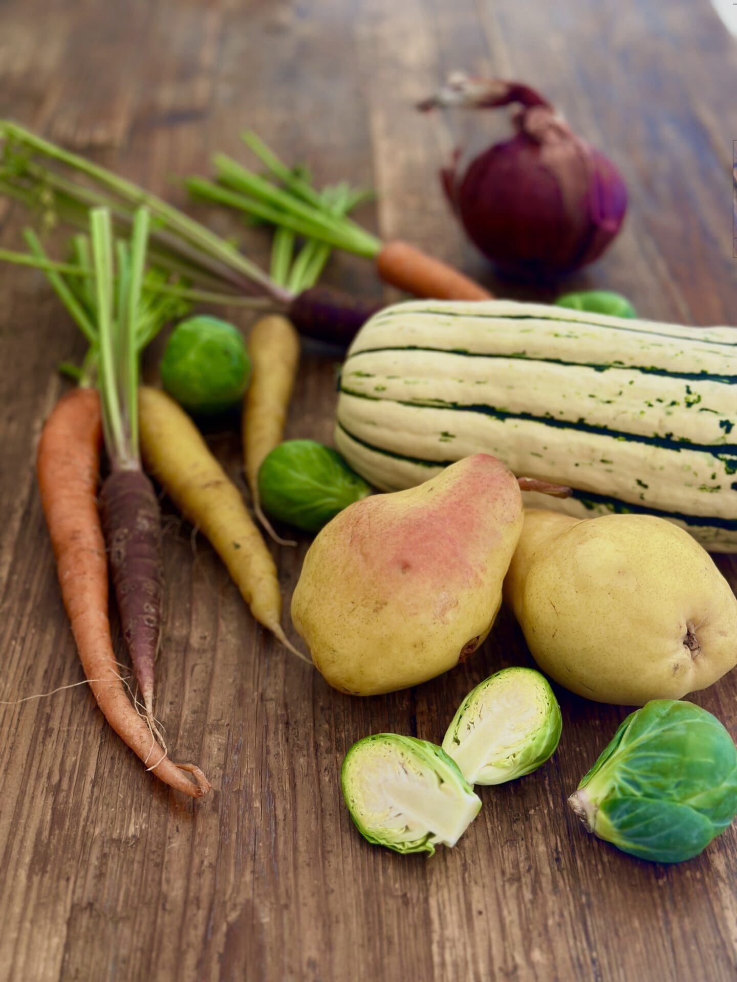 A selection of colourful carrots, Brussel sprouts, pears, Delicata squash and red onion sits on a wood harvest table ready to be made into a Thanksgiving side dish.
