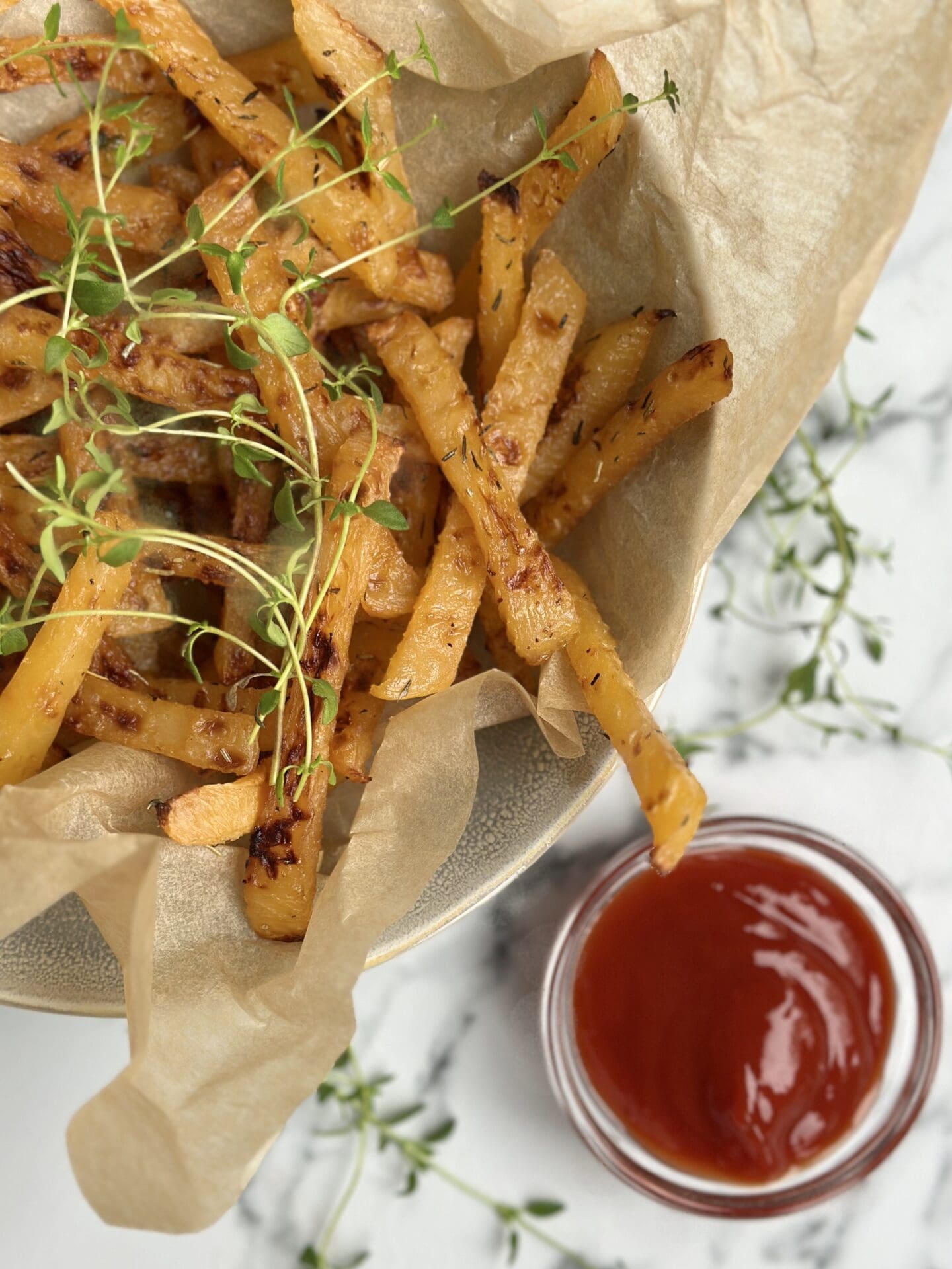 IMG_9701 A brown paper lined bowl of golden rutabaga fries garnished with fresh thyme sits on a white marble counter top along with a small bowl of red ketchup