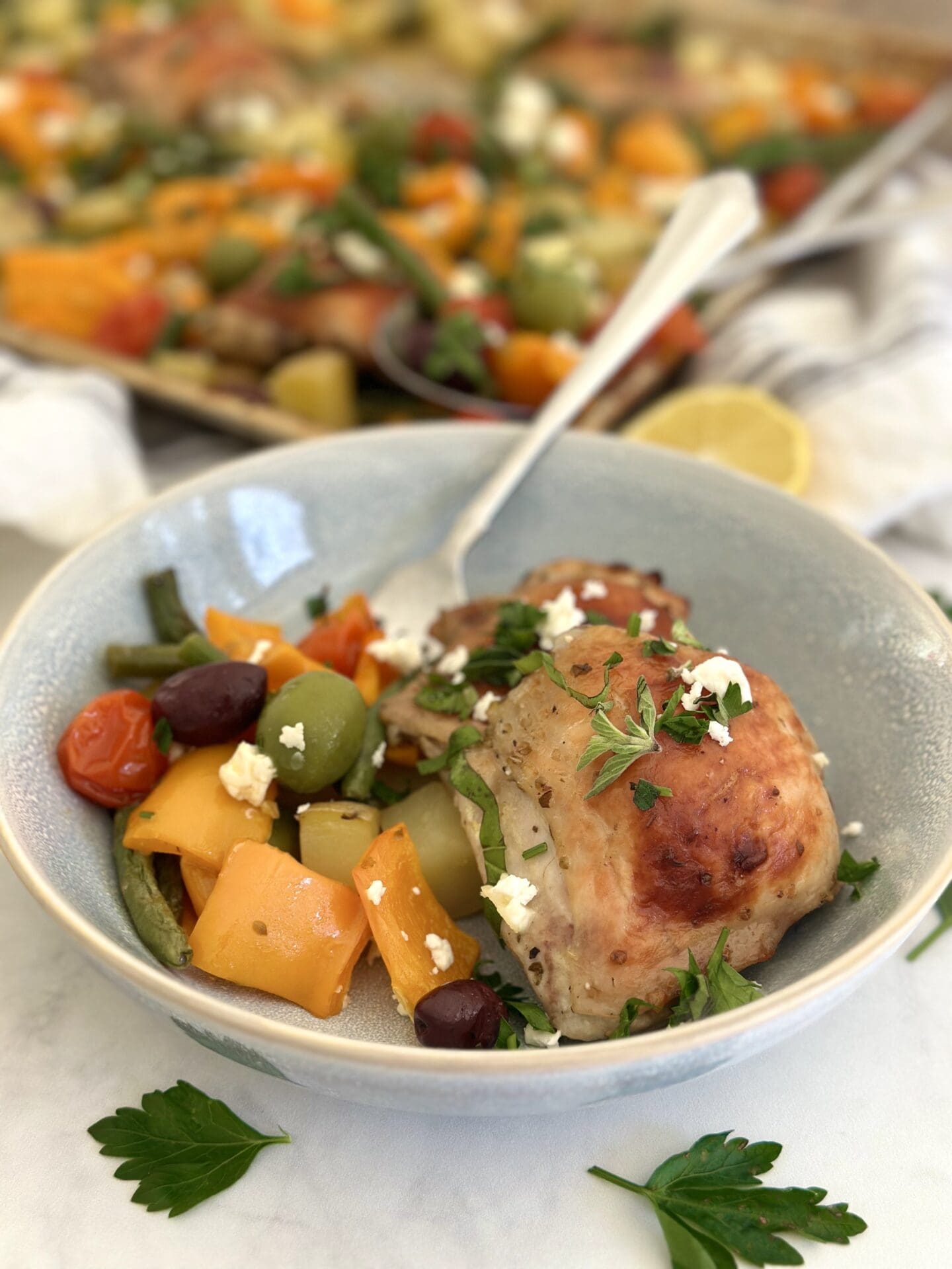 Greek Sheet Pan Chicken and Vegetables A bowl of Greek Chicken Sheet Pan Dinner is seen with golden chicken thighs, roasted vegetables, olives and feta cheese. The sheet pan with the rest of the dinner is seen in the background.