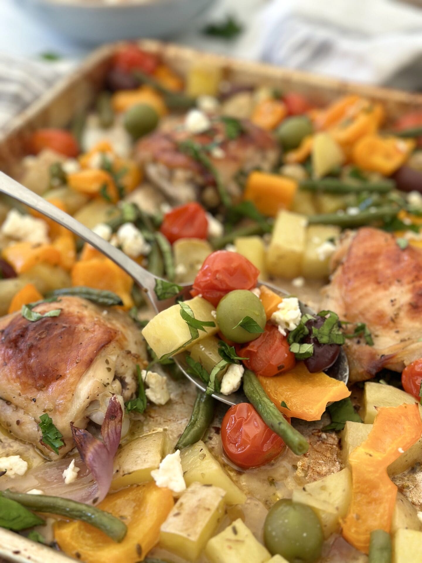 A large spoon scoops roasted vegetables, olives, fresh herbs and feta cheese from a large tray of Greek Chicken Sheet Pan Dinner
