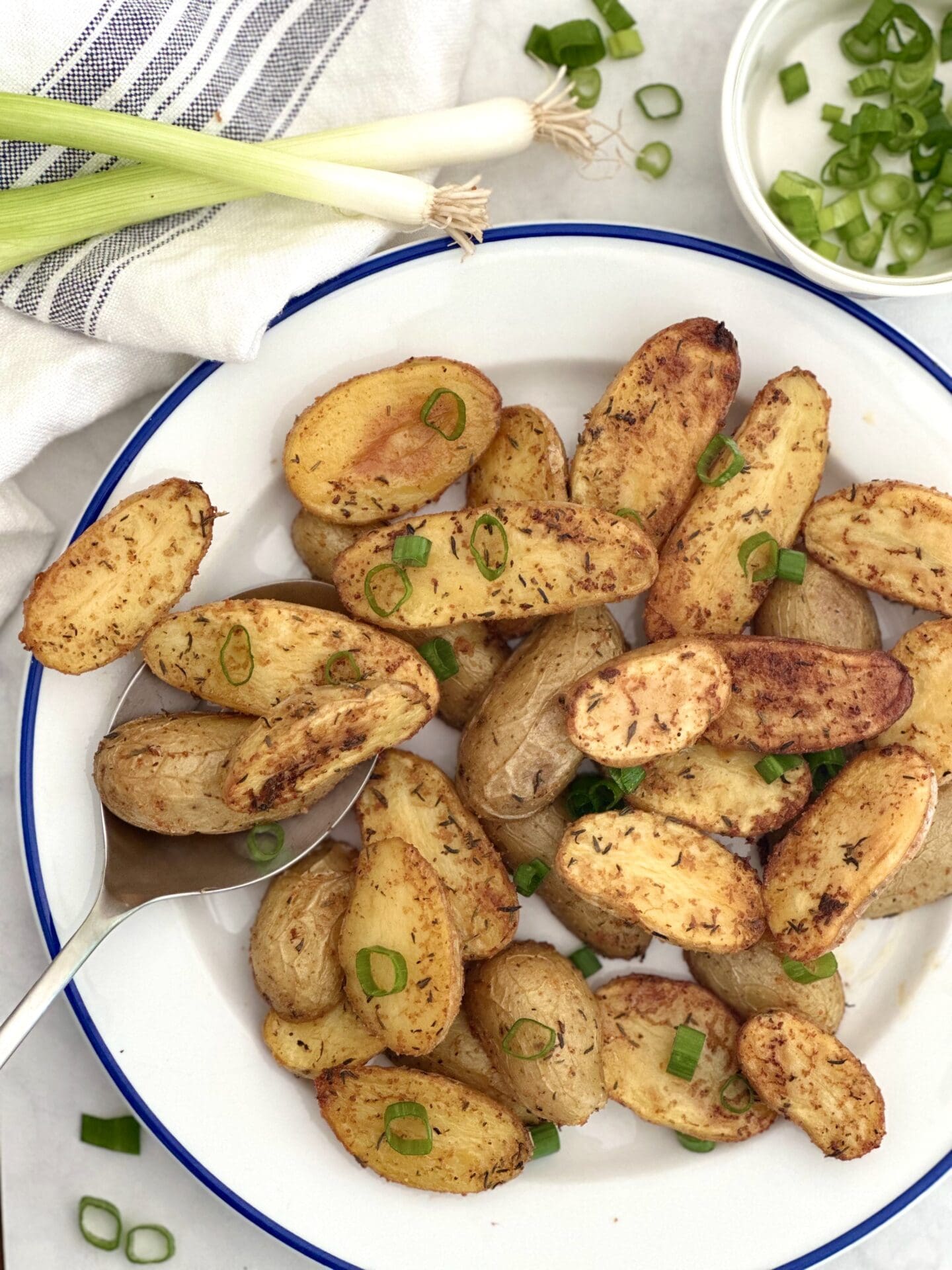 A blue trimmed plate of golden roasted fingerling potatoes is seen from above, surrounded by green onions