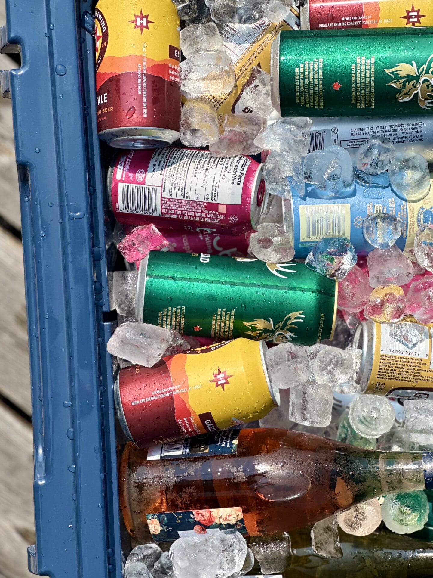 A well packed, sun lit cooler filled with ice, cans and bottles is seen from above, sitting on a wood dock