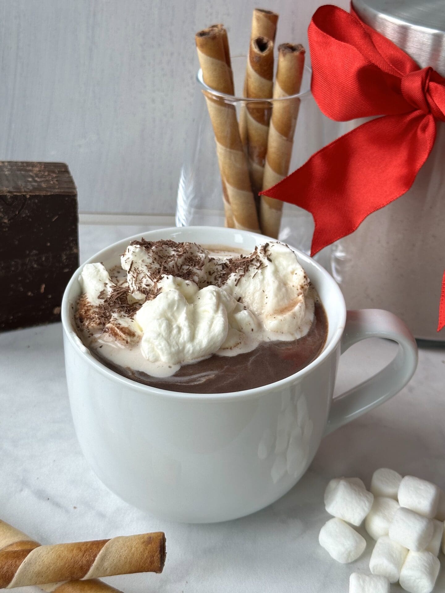 A large mug filled with whipped cream-topped hot chocolate is surrounded by cookie rolls, marshmallows and a big block of chocolate. A jar of the hot chocolate mix, tied up with a festive red bow for gifting is seen in the background.