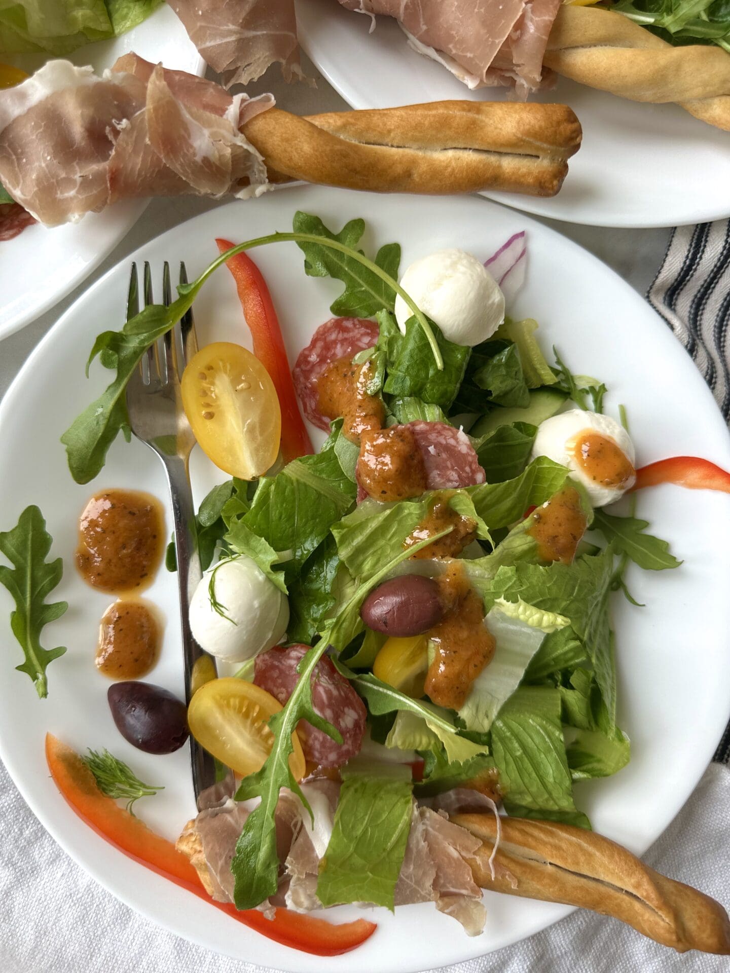 IMG_3753 A single serving of Antipasti Salad with Prosciutto Wrapped Breadsticks is seen from above, drizzled in sundried tomato vinaigrette. More breadsticks can be seen in the background.