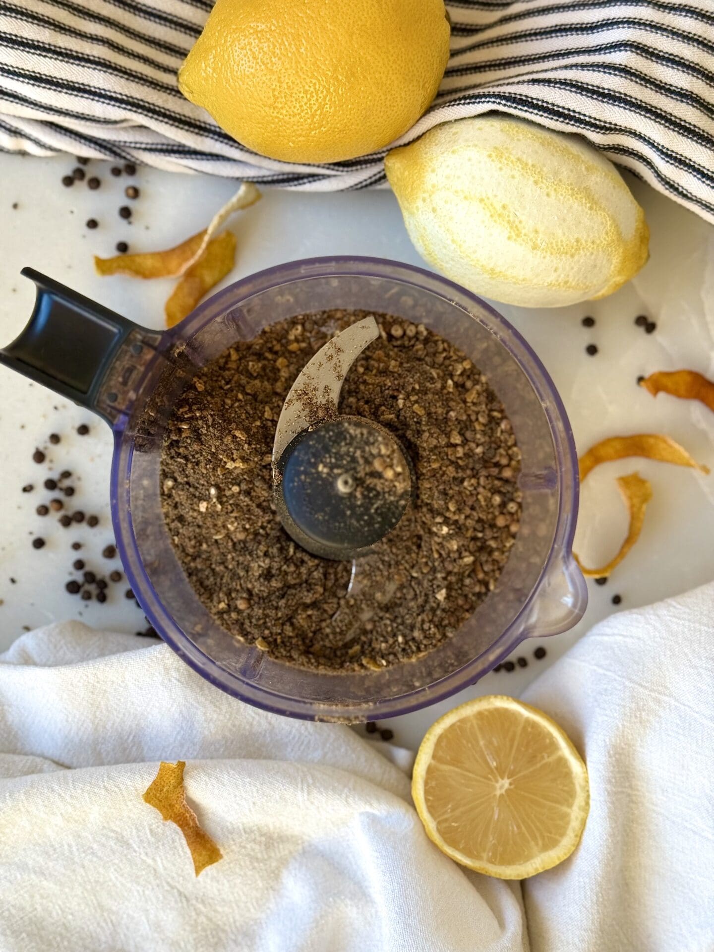 IMG_4126 A mini food processor is seen from above, containing Homemade Lemon Pepper Seasoning. The appliance is surrounded by fresh lemons, lemon zest and peppercorns.