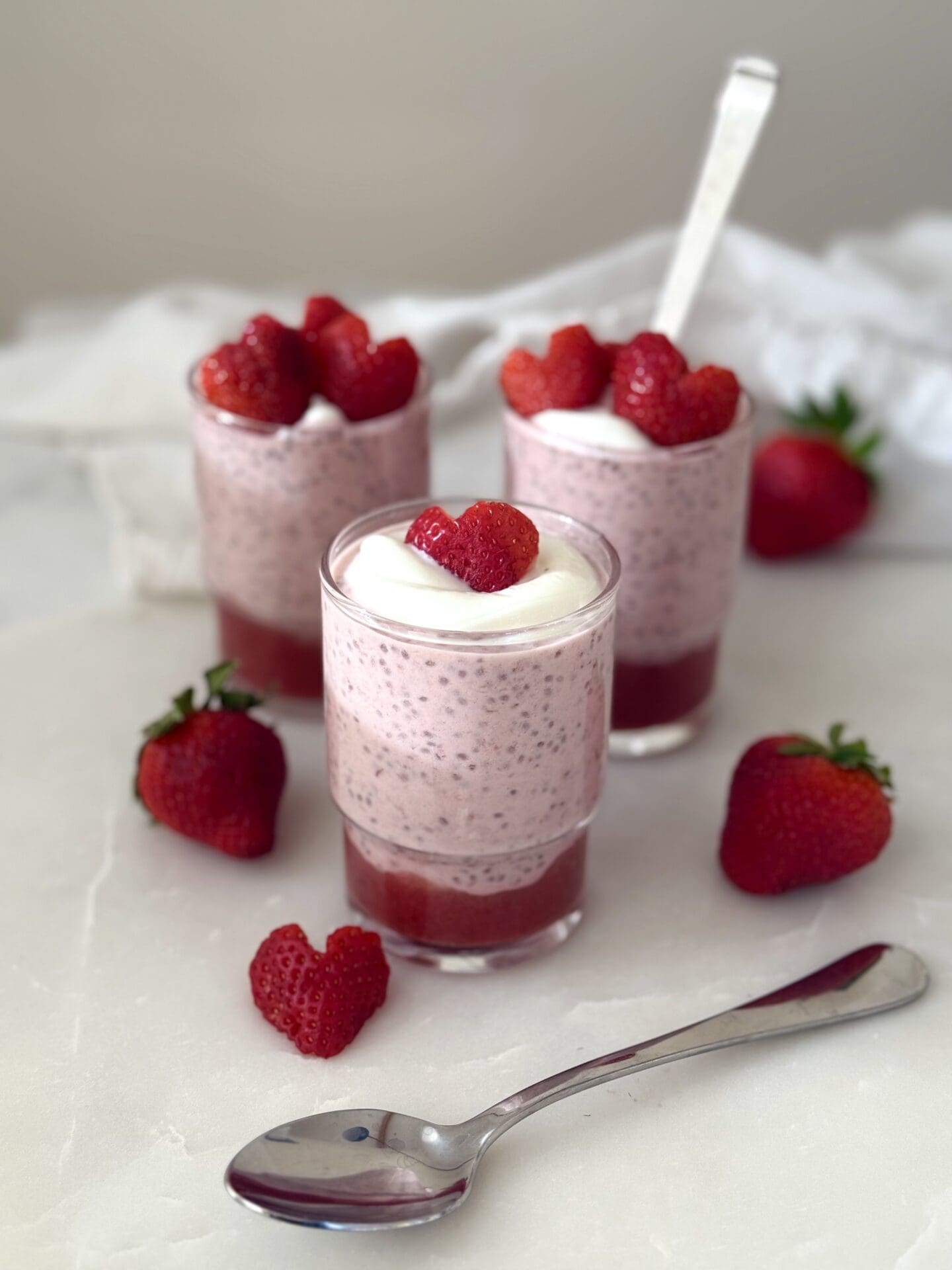 A trio of glass jars hold individual servings of strawberry chia seed pudding. The puddings are garnished with heart shaped strawberry slices.