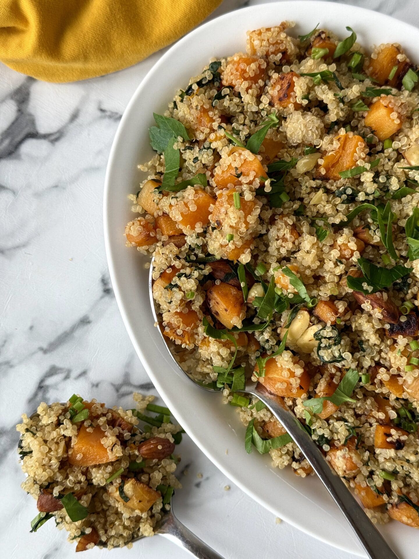 Half of a serving platter full of quinoa salad with butternut squash, spinach and crunchy almonds is seen on white marble counter. A serving spoon overflowing with the salad sits beside the platter.