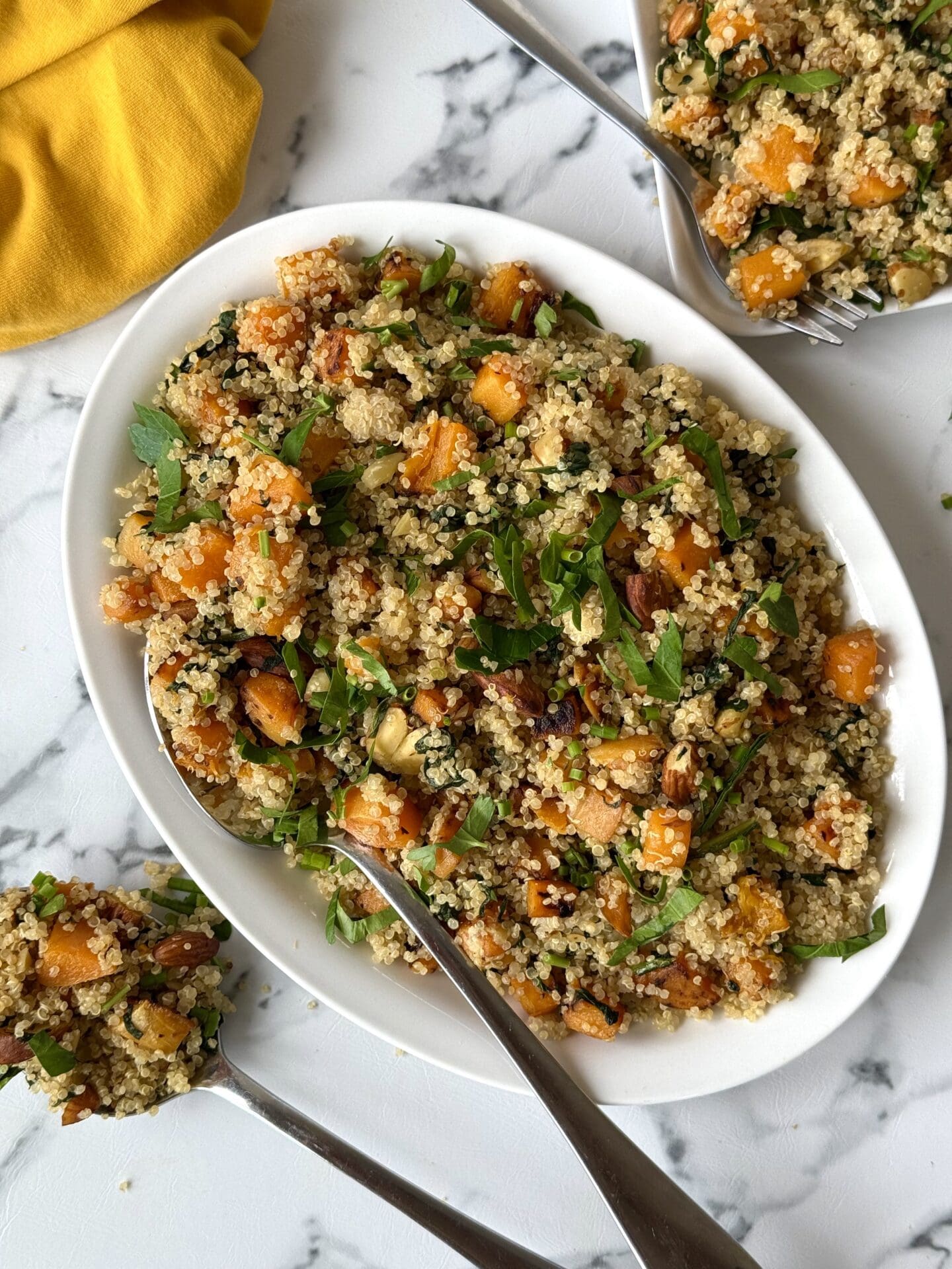 A platter of quinoa salad with butternut squash, spinach and crunchy almonds is seen from above on a white marble counter, surrounded by serving utensils and a yellow dinner napkin.