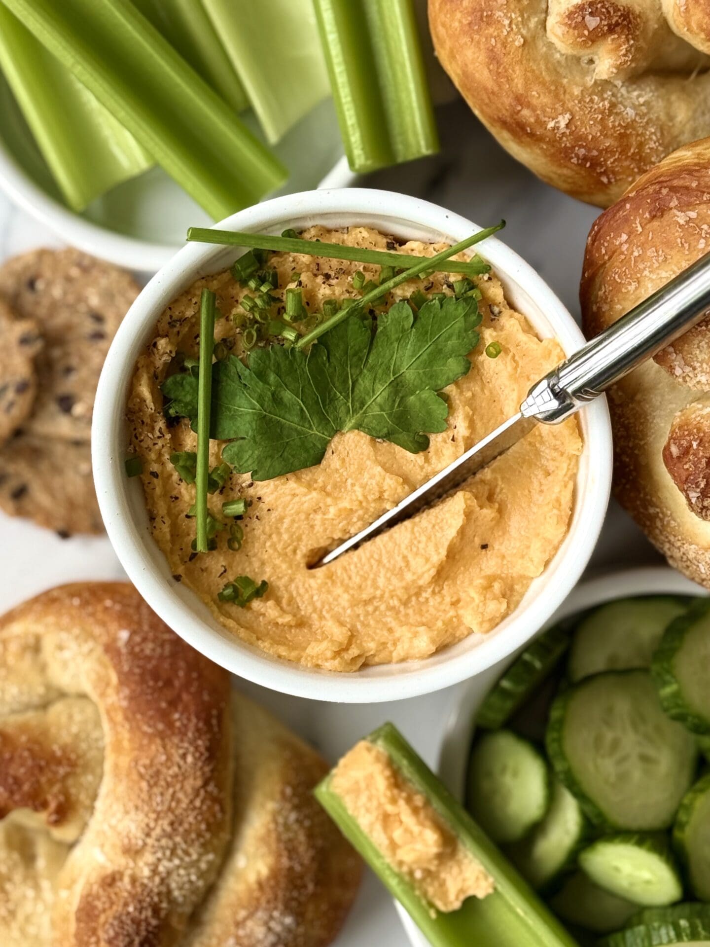 A dish of Pub Style Beer Cheese is seen from above, garnished with fresh herbs and surrounded by green vegetables and soft pretzels.
