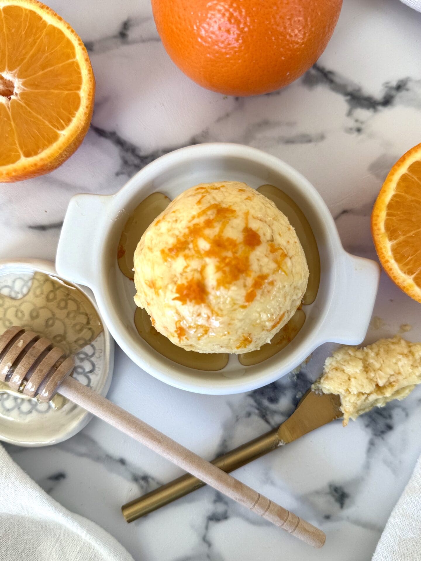 A sphere of Orange and Honey Butter is seen from above. drizzled with honey and sprinkled with fresh orange zest. The butter rests in a dish on a white marble counter, surrounded by fresh oranges, a butter spreader and a honey dipper.