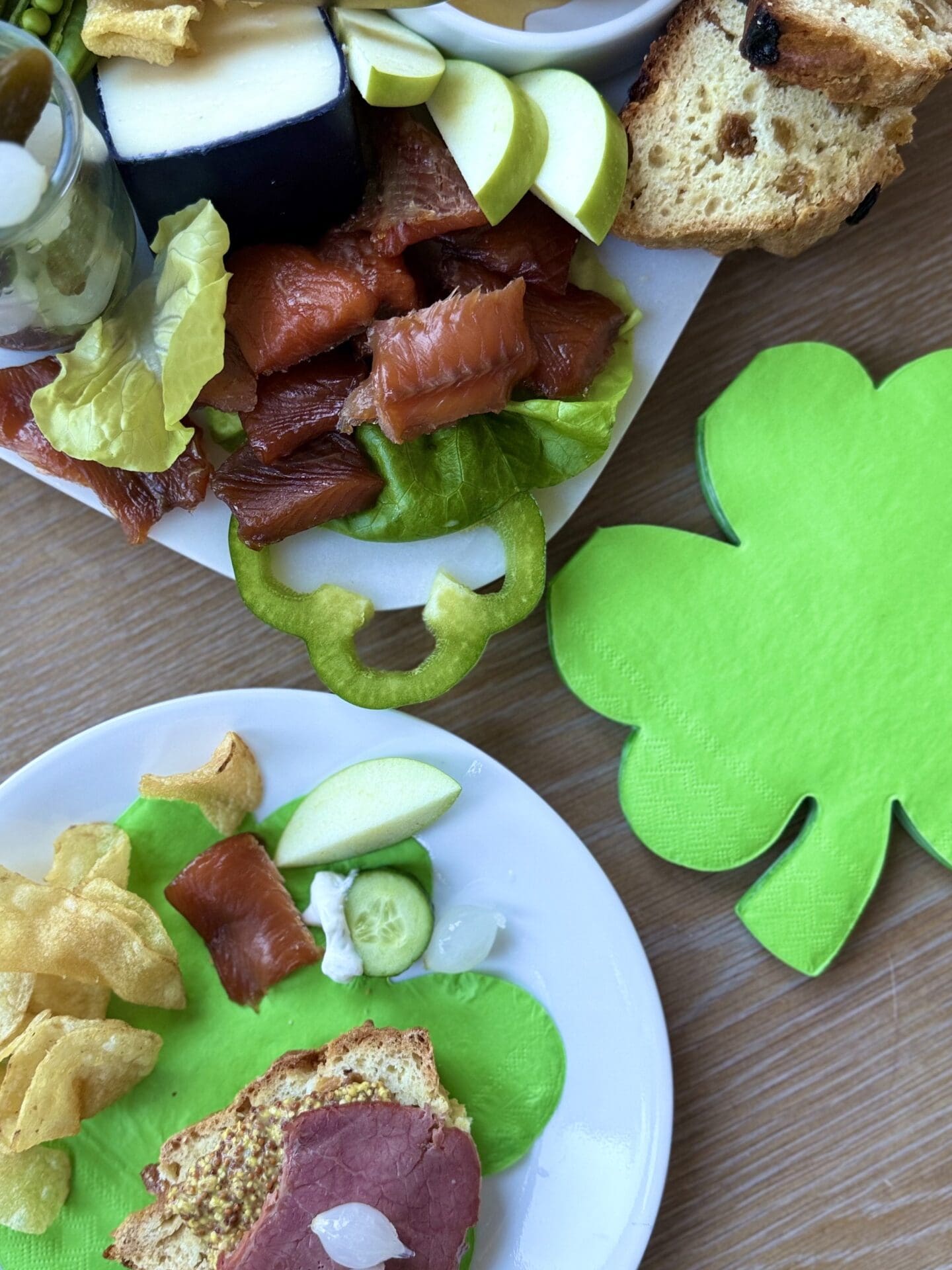 A St Patrick's Day Charcuterie Board is seen along with an individual plate containing a selection of items from the board. A green shamrock shaped napkin is seen to the side of the photo.