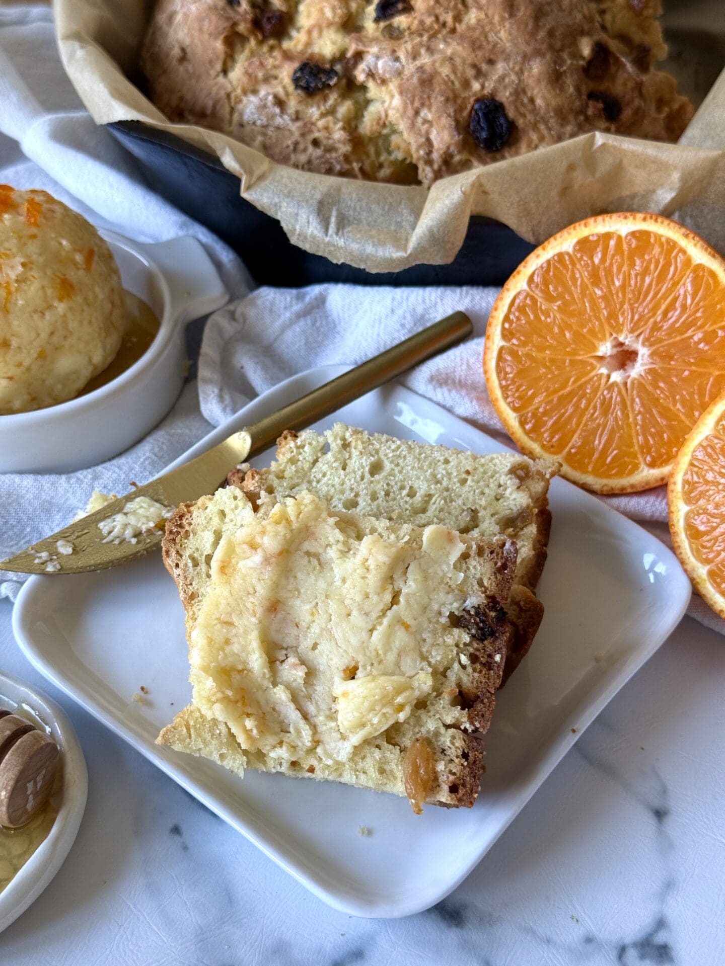 Homemade Orange and Honey Butter is slathered on slices of freshly baked Irish soda bread. A loaf of the soda bread can be seen in a cast iron skillet in the background. Fresh oranges and a honey dipper complete the image.