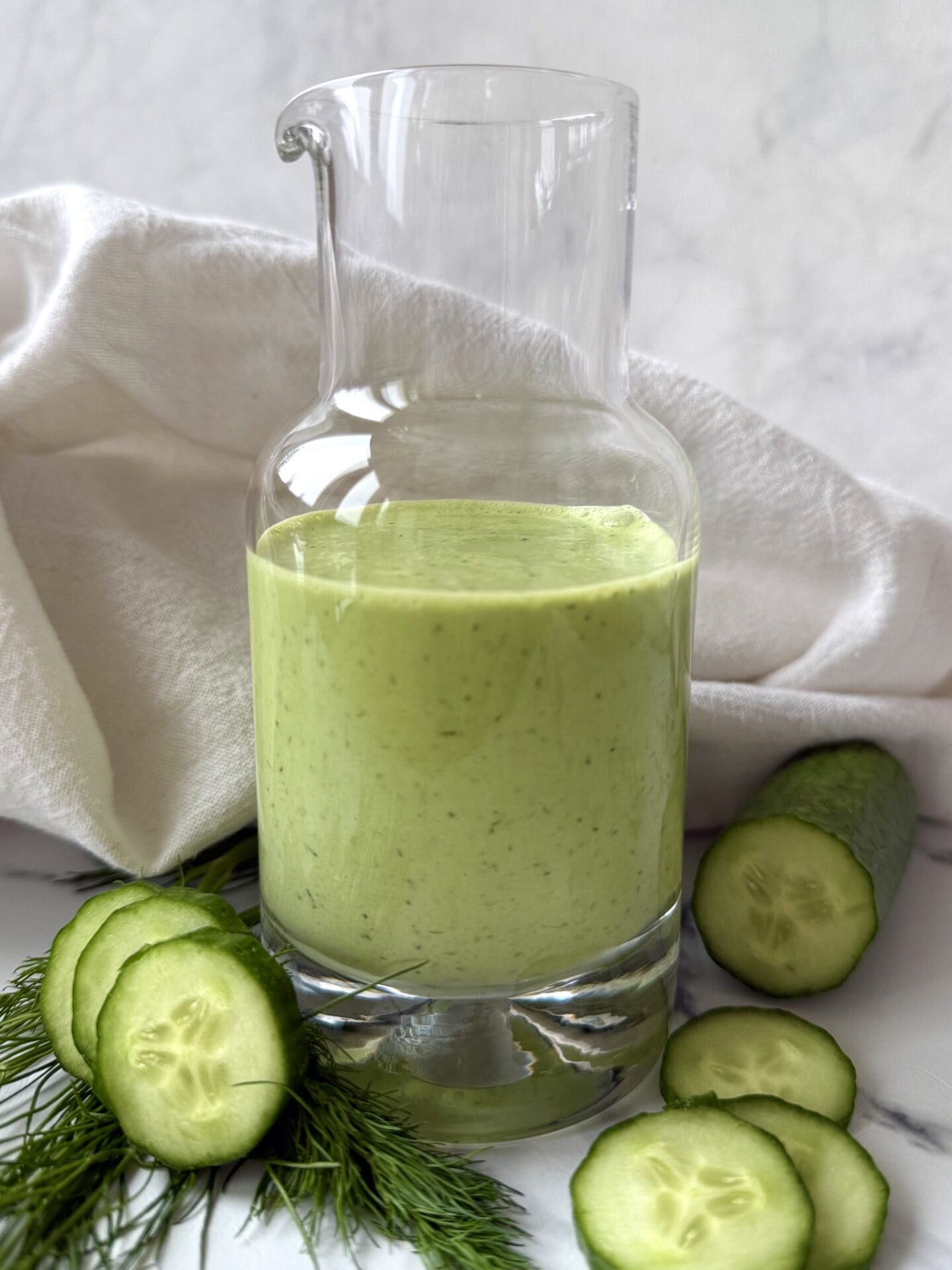 IMG_4924 A glass bottle of Creamy Cucumber Dill Salad Dressing is seen on a white background, surrounded by fresh dill and sliced cucumbers