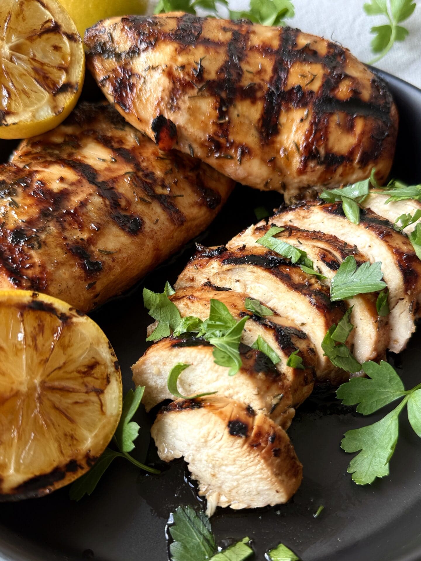 Sweet and Tangy Marinated Grilled Chicken is seen on a black platter with grilled lemon wedges and fresh parsley.