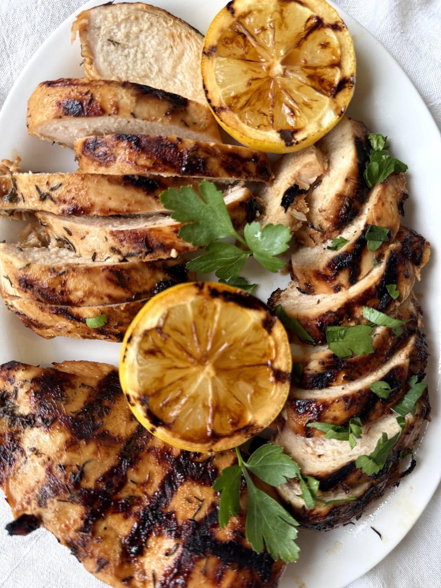 A white platter of sweet and tangy marinated grilled chicken is seen from above, on a white linen tablecloth. The chicken is garnished with grilled lemon and fresh parsley.