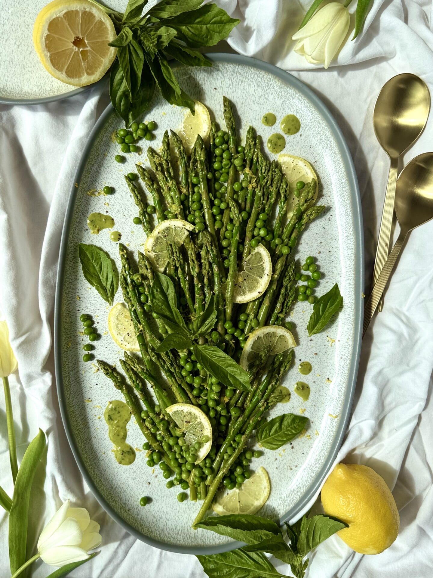 A large platter of Lemon Basil Asparagus and Peas rests in the centre of the sunlit image. Fresh basil, lemons, gold serving spoons and pale yellow tulips complete the image.