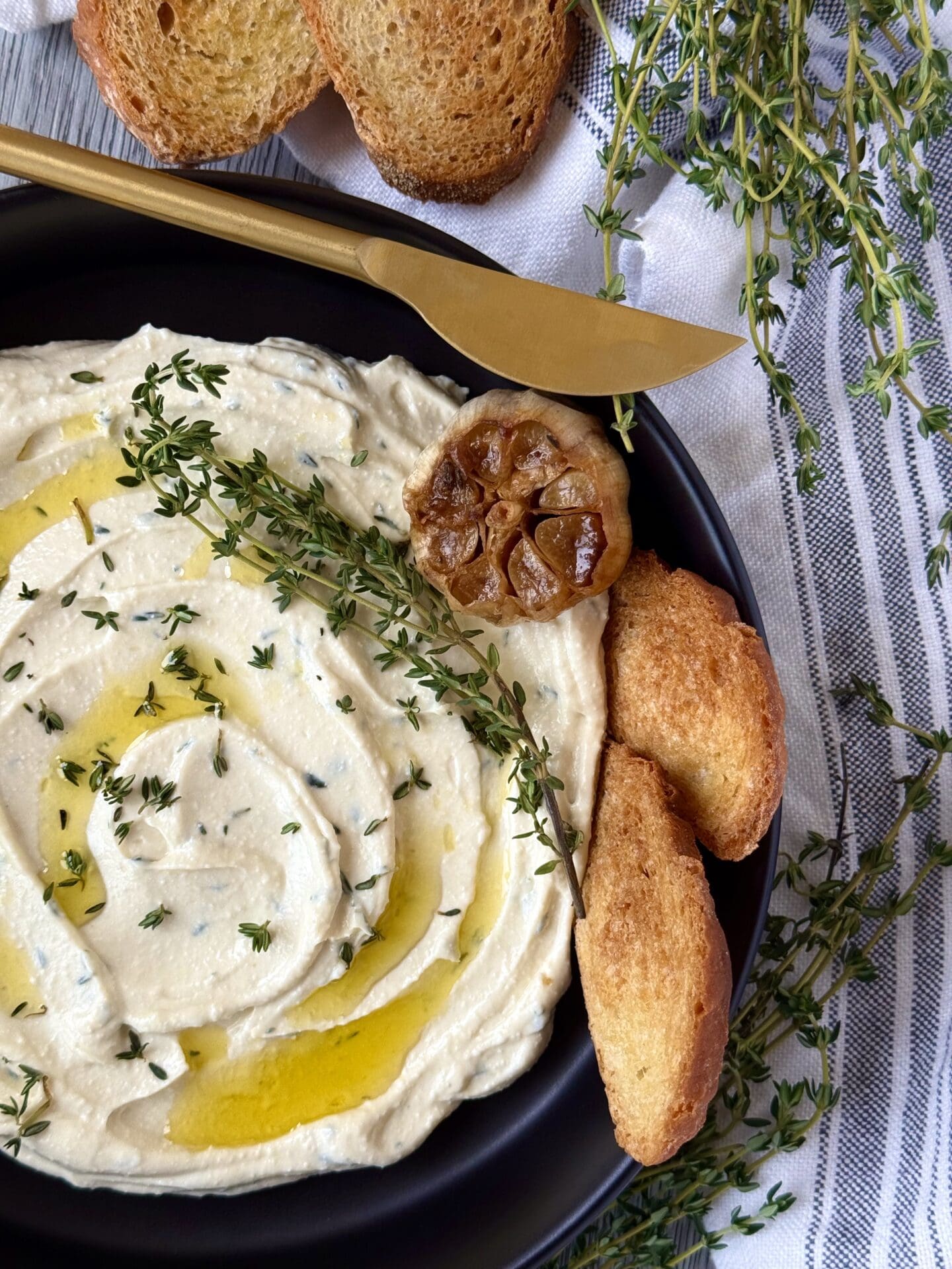 A black bowl filled with swirled Whipped Feta with Roasted Garlic and Thyme dip is seen on a grey wood table. The dip is garnished with drizzles of olive oil, a head of roasted garlic and sprigs of fresh thyme. The dish is surrounded by slices of toasted baguette and sprigs of fresh thyme. A gold spreader sits on the edge of the dish.
