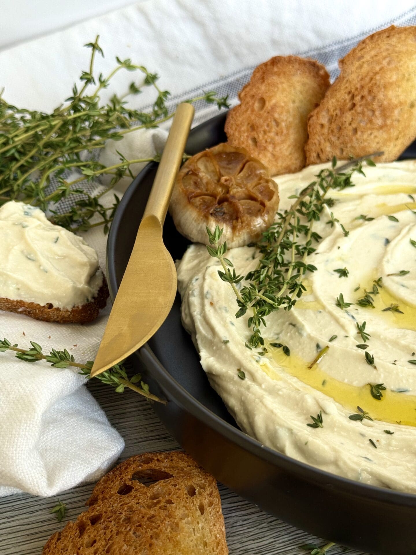 Half of a black dish filled with Whipped Feta with Roasted Garlic and Thyme dip fills the right side of the image. The dip is lavishly garnished with olive oil and fresh thyme. A white and grey striped kitchen towel surrounds the bowl and sprigs of fresh thyme and toasted baguette slices complete the image.