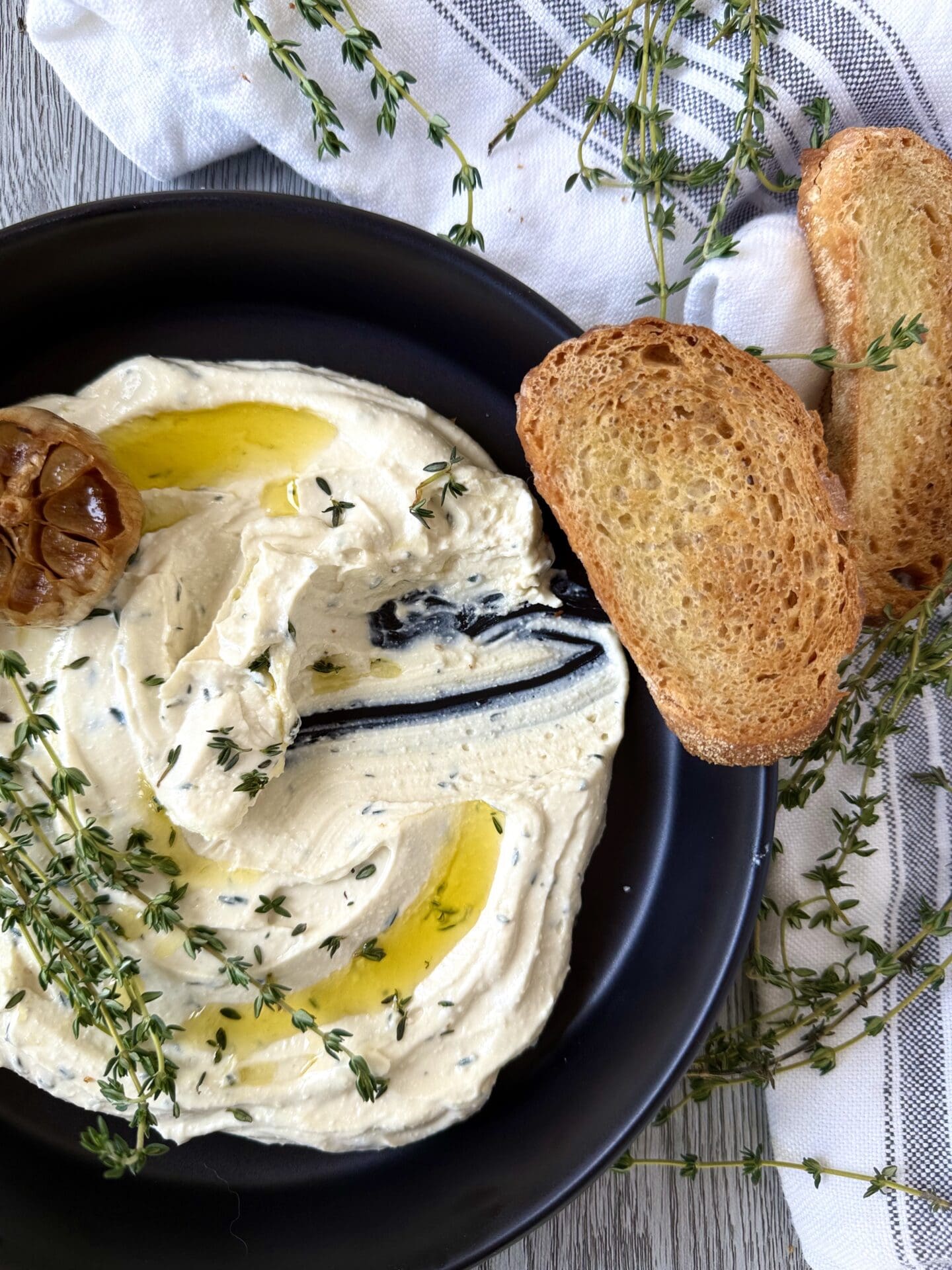 A bowl of Whipped Feta with Roasted Garlic and Thyme is seen from above. A swoop of dip is missing, implying that someone has used one of the slices of golden toasted baguette to scoop up the dip.