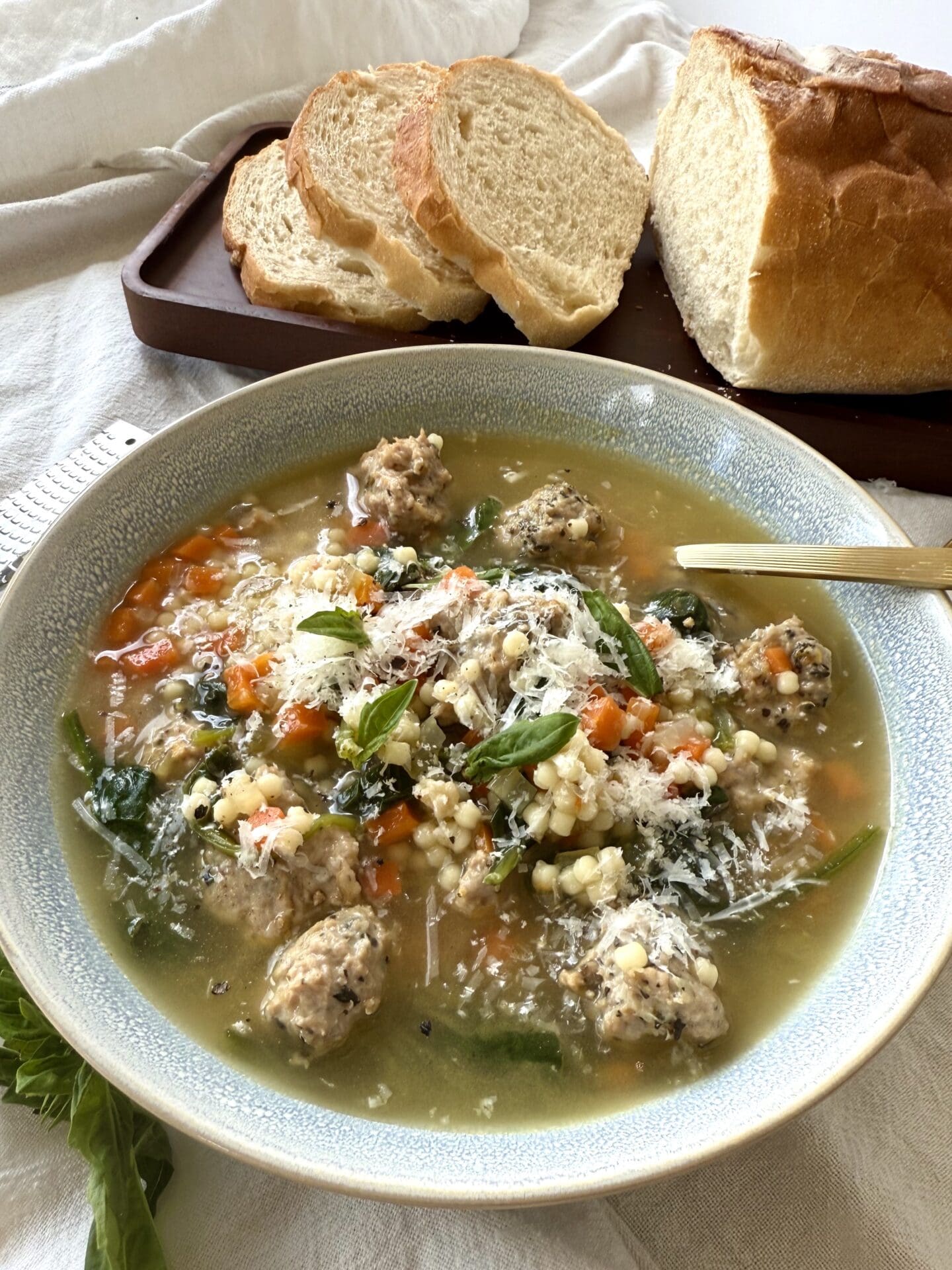 IMG_5720 A bowl of Healthier Italian Wedding Soup sits on a table, laden with roasted turkey meatballs and garnished with freshly grated pecorino Romano cheese and basil leaves. A loaf of fresh Italian bread sits in the background.