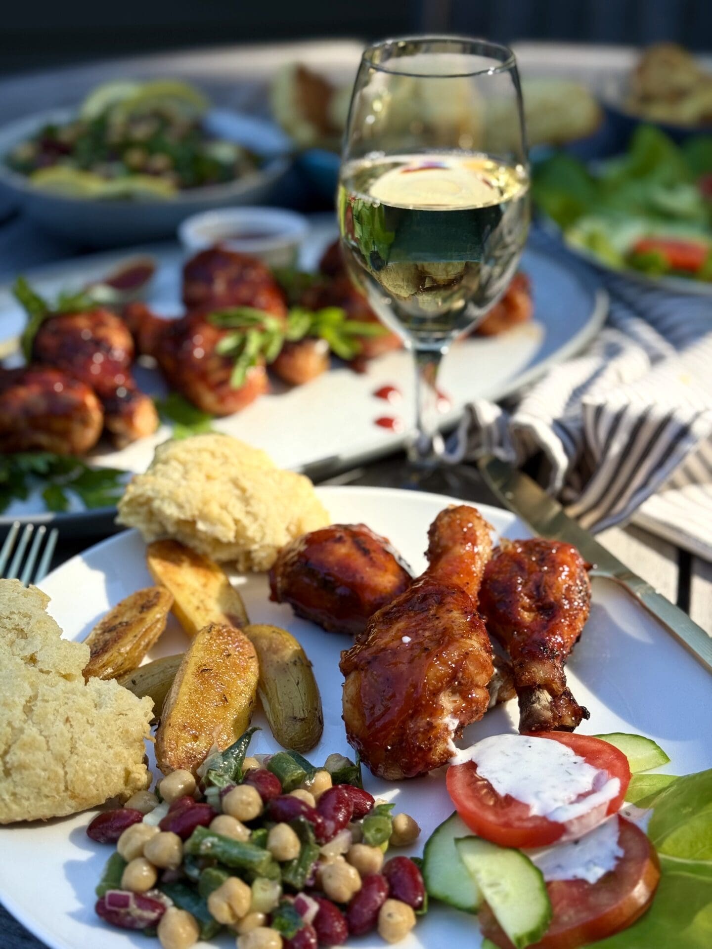 IMG_5960 An outdoor table filled with platters of Smokey Sweet Grilled Chicken Legs, bean salad and other side dishes is seen in the fading evening sunlight. A glass of white wine sits in the centre of the table catching the light.