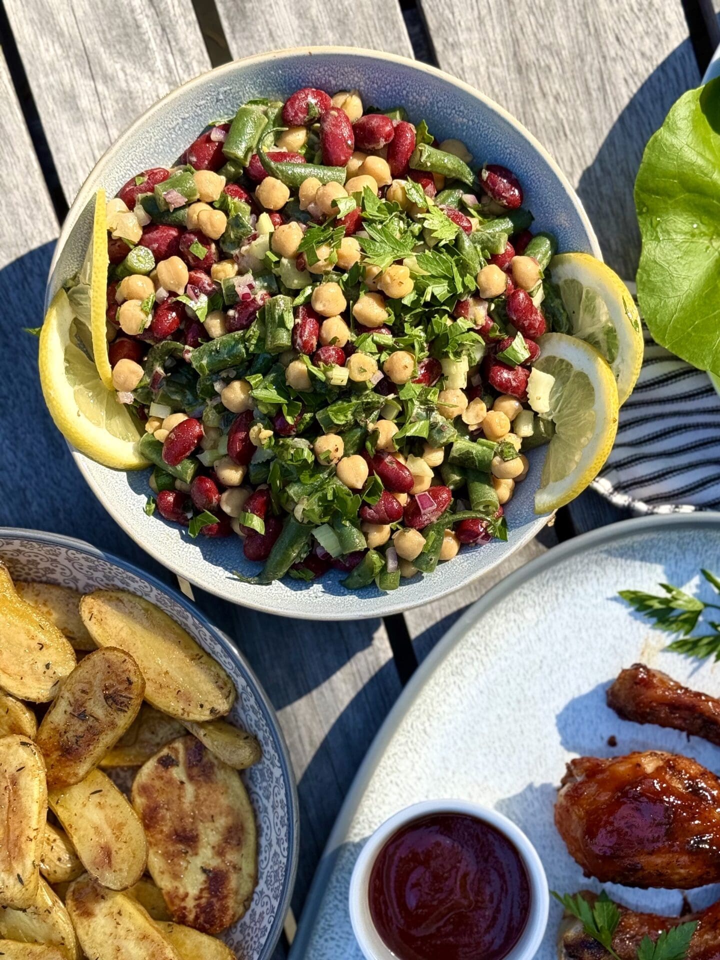 A bowl of Marinated Mediterranean Bean Salad is seen from above on an outdoor table, as part of a backyard barbecue dinner. The salad is surrounded by platters of grilled chicken and roasted potatoes.