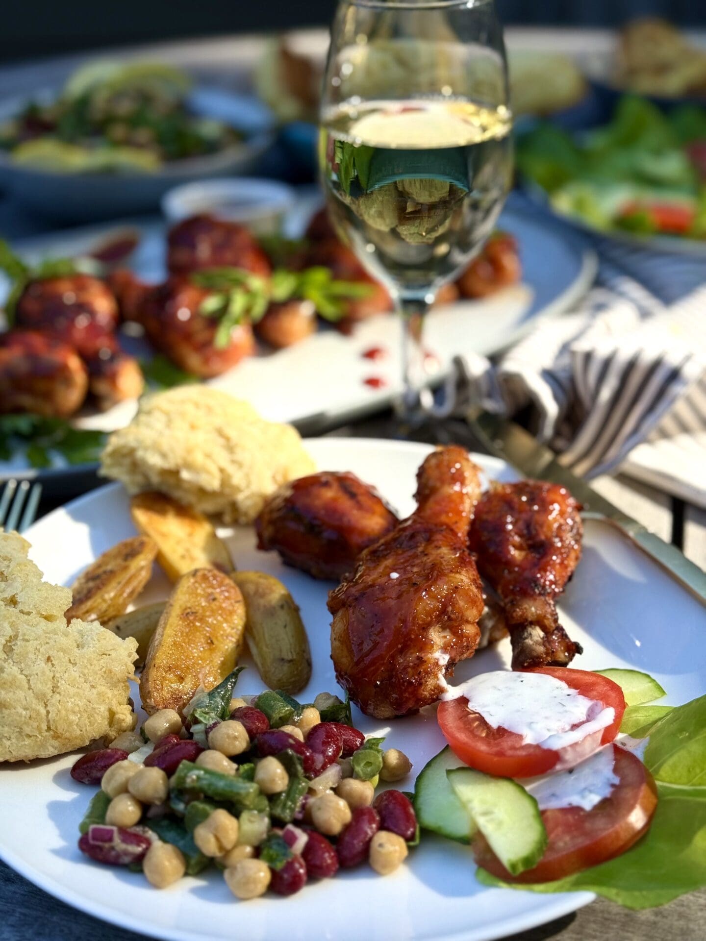A dinner plate of Marinated Mediterranean Bean Salad with roasted potatoes, salad and grilled chicken is seen on a picnic table. A glass of cold white wine and platters of food are seen in the background.