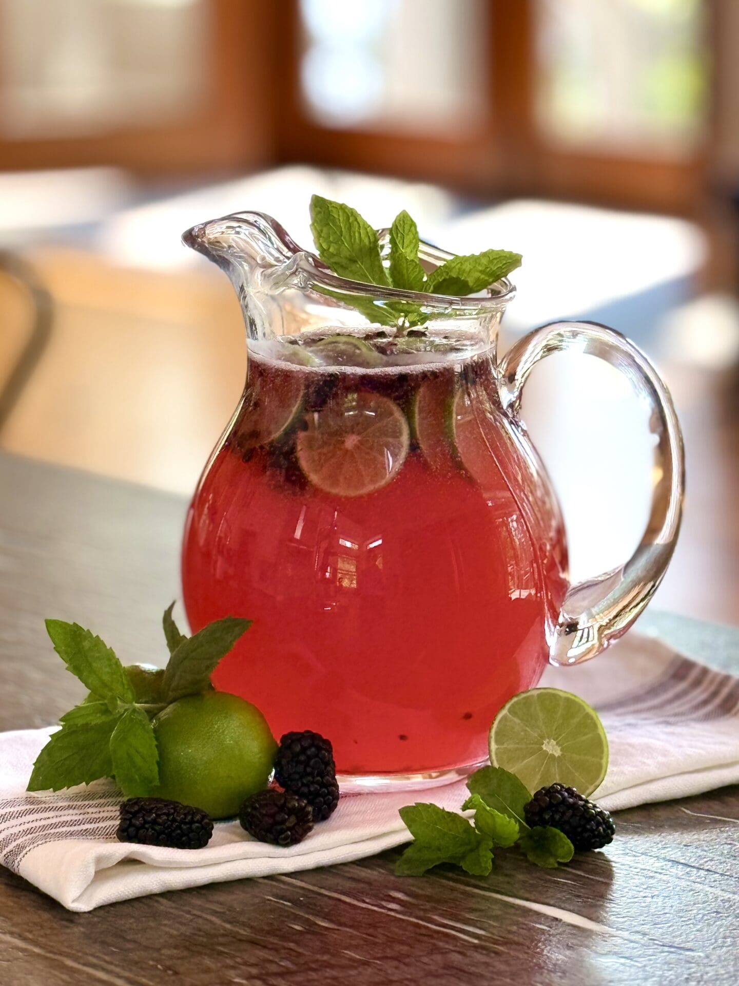 A glass pitcher full of Blackberry Mojito Mocktail sits on a table, garnished with blackberries, lime wedges and fresh mint. Sunshine streams in through the screen doors in the background.