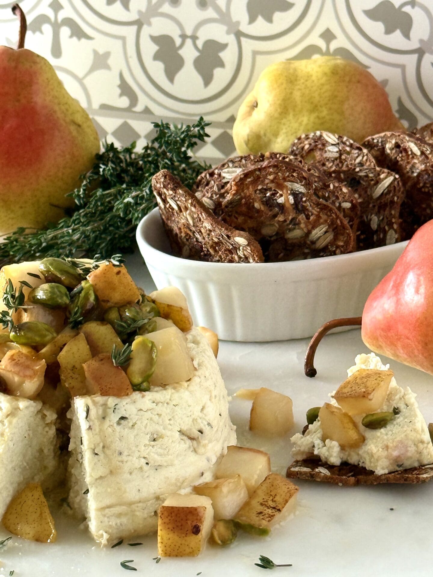 IMG_6965 Pistachio and Pear Baked Ricotta is shown, lavishly garnished with caramelized pears, green pistachios and sprigs of fresh time. The appetizer is surrounded by crisp crackers for serving and red and yellow pears.