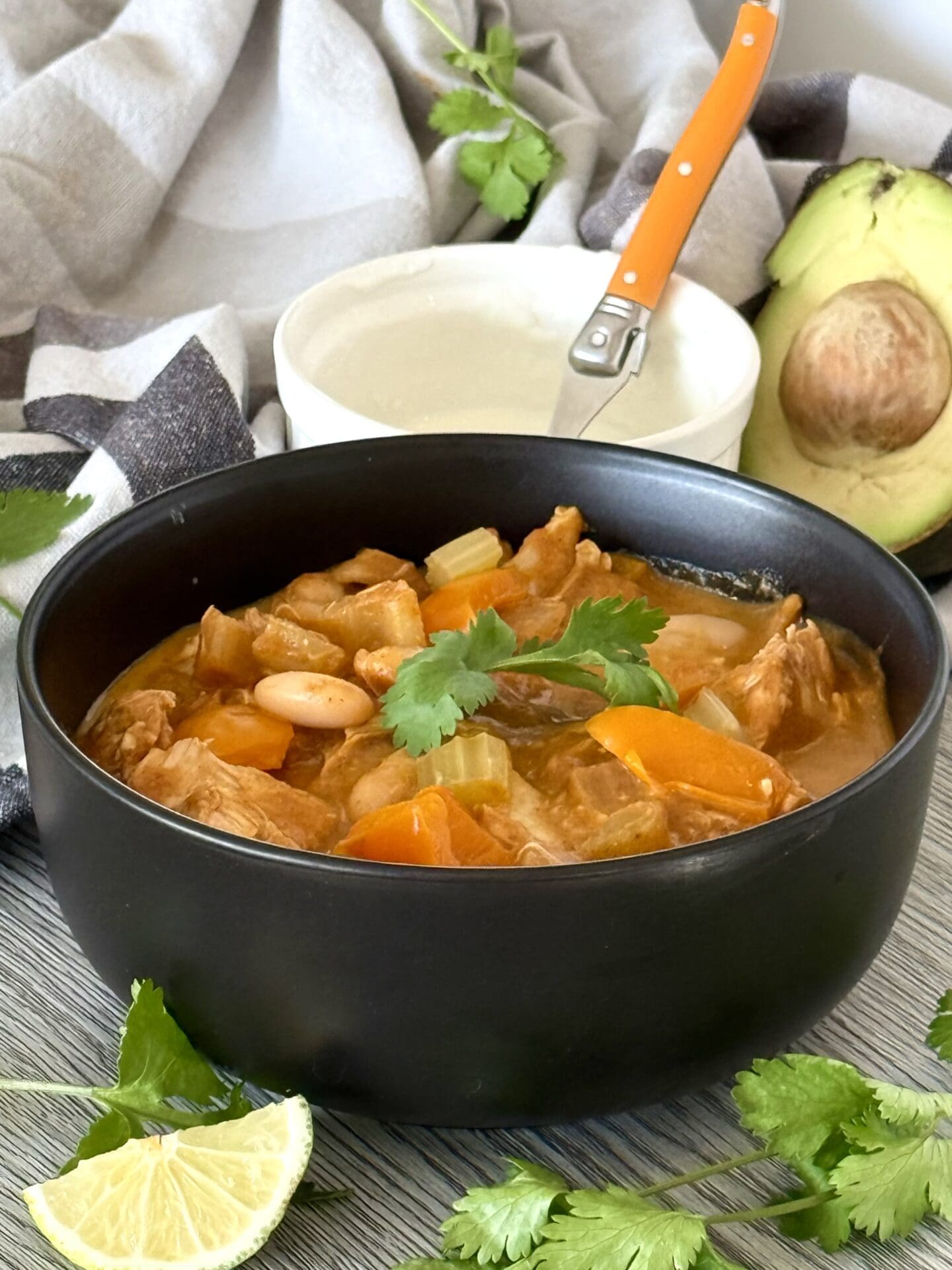 IMG_7241 A black bowl of Pumpkin Chicken Chili sits on a table, surrounded by fresh avocado, cilantro, lime wedges and a dish of sour cream.