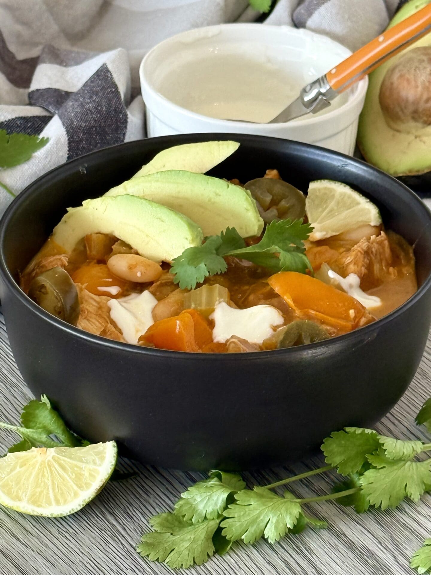IMG_7247 A black bowl filled with garnished Pumpkin Chicken Chili sits on a grey wood table. A bowl of sour cream and a avocado half are seen in the background.