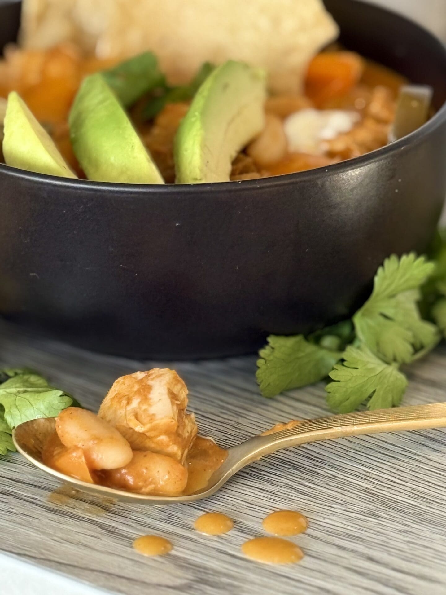 IMG_7280 A spoonful of Pumpkin Chicken Chili rests on a table, surrounded by drips of its delicious orange hued sauce. A black bowl full of lavishly garnished chili sits in the background, overflowing with fresh avocado, cilantro and corn chips.