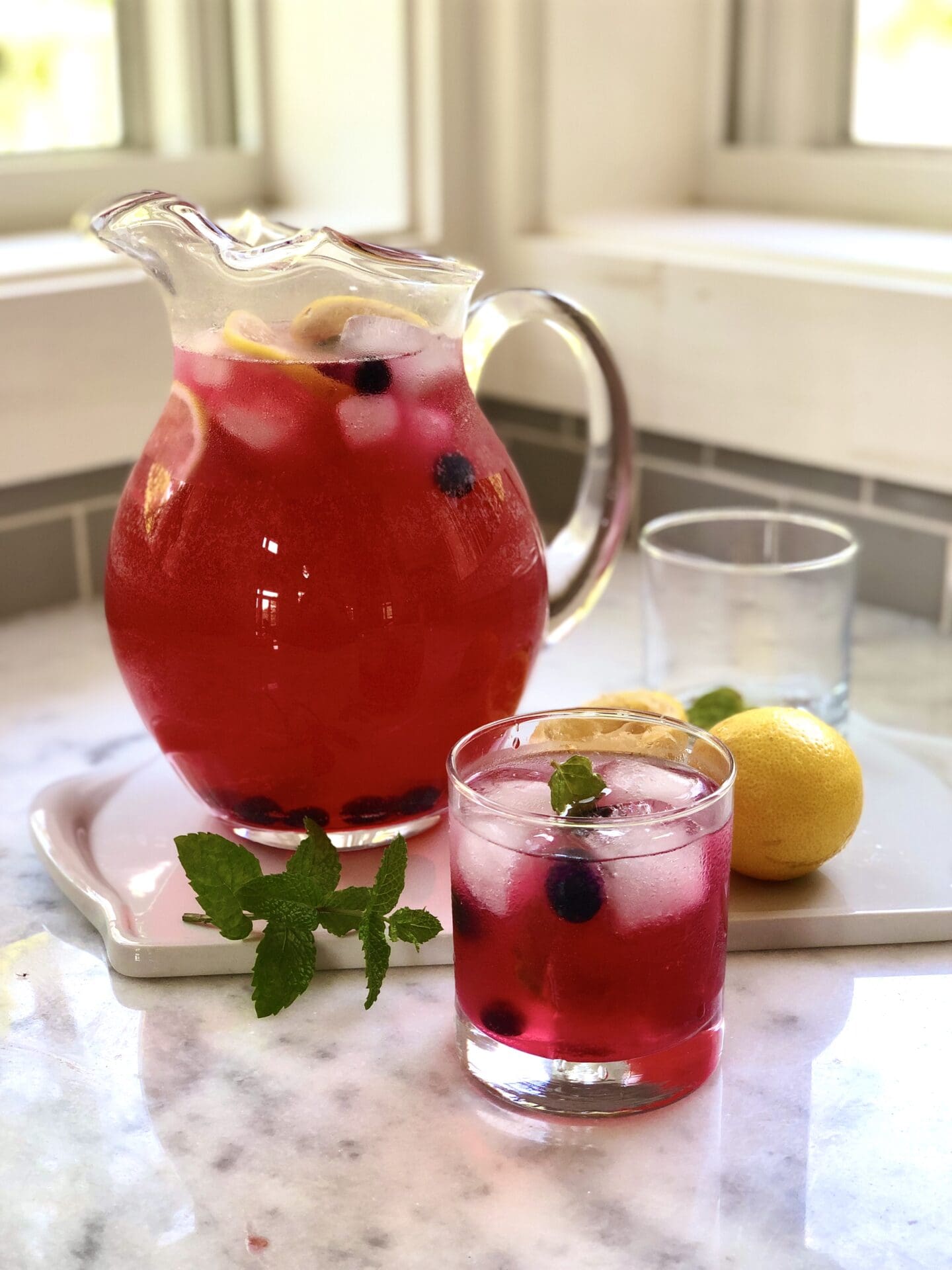 IMG_4797 A pitcher of ice cold blueberry lavender lemonade sits on a white marble counter by a window. A glass of the drink sits in the foreground, garnished with lemon slices and fresh blueberries.