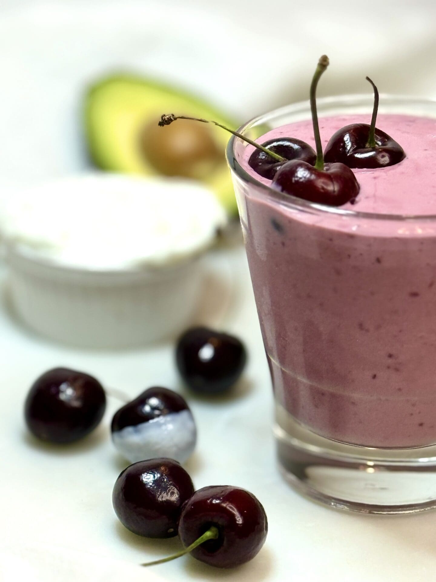 A glass of pink cherry vanilla smoothie slides into the frame from the side, surrounded by fresh cherries, yogurt and avocado.