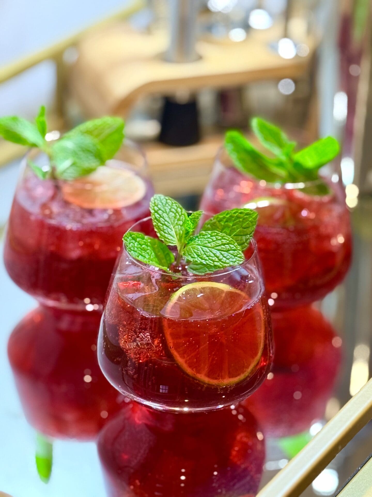 IMG_7784 Three glasses of Pomegranate Lime Mocktail are garnished with fresh mint, lime wheels and pomegranate arils. The glasses sit together on a mirrored bar cart, which shows their reflection.