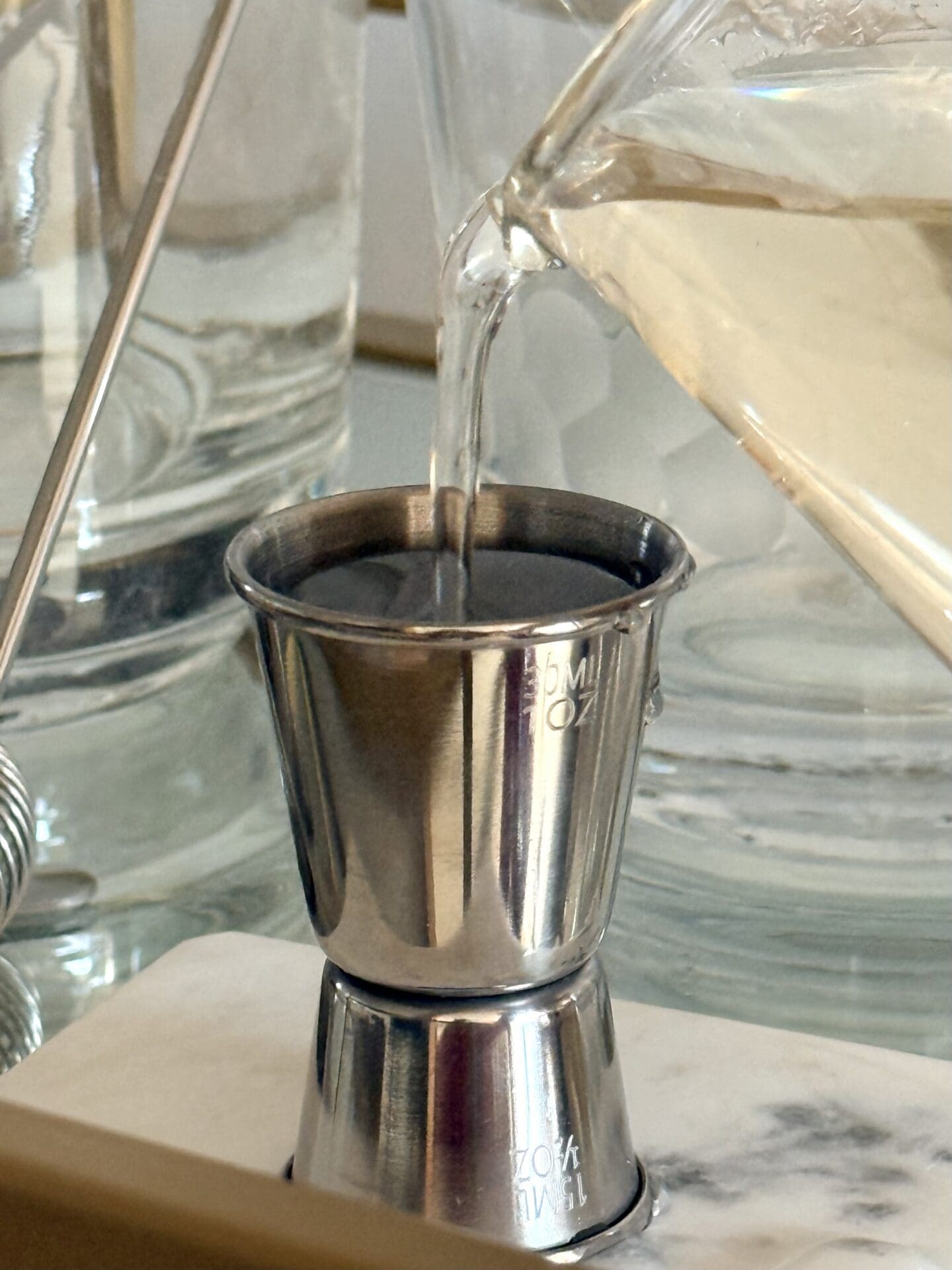 Homemade simple syrup pours from a glass beaker into a silver jigger.