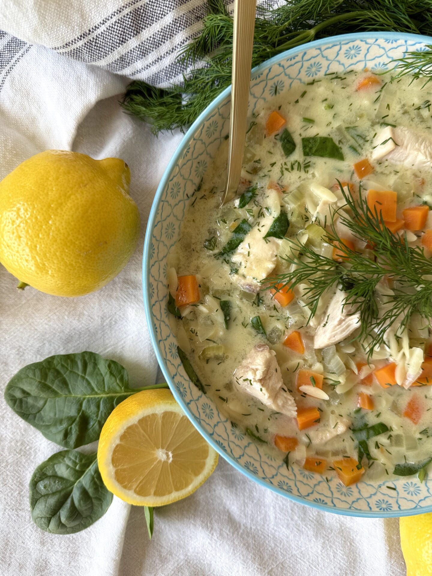 IMG_5278 A bowl of Avgolemono soup is seen entering the frame from the right so half of the bowl can be seen in the image. The soup is surrounded by lemons and baby spinach leaves.