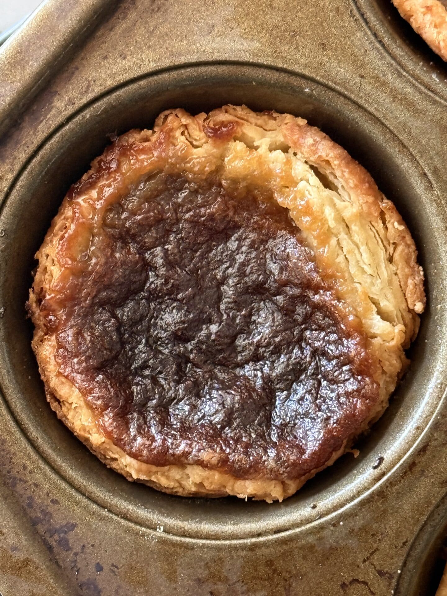 IMG_8428 A single butter tarts is seen from above, sitting in its pan. The flaky, golden layers of the pastry crust can clearly be seen along with the glossy, caramel-like filling.