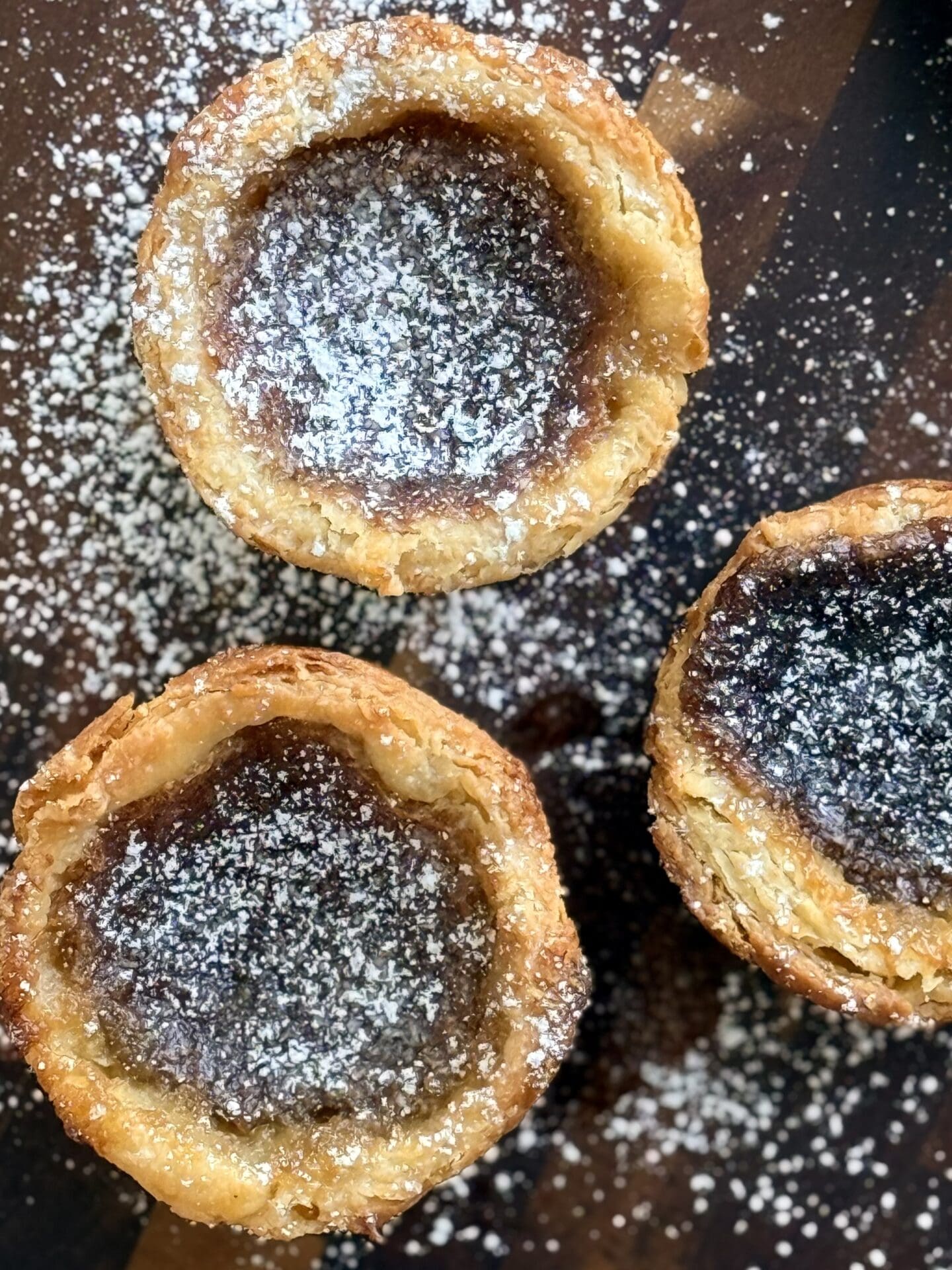 IMG_8439 A trio of freshly baked butter tarts are seen from above, lightly dusted with powdered sugar. The butter tarts show off the flaky, golden layers of their crusts and their silky, caramel-like filling