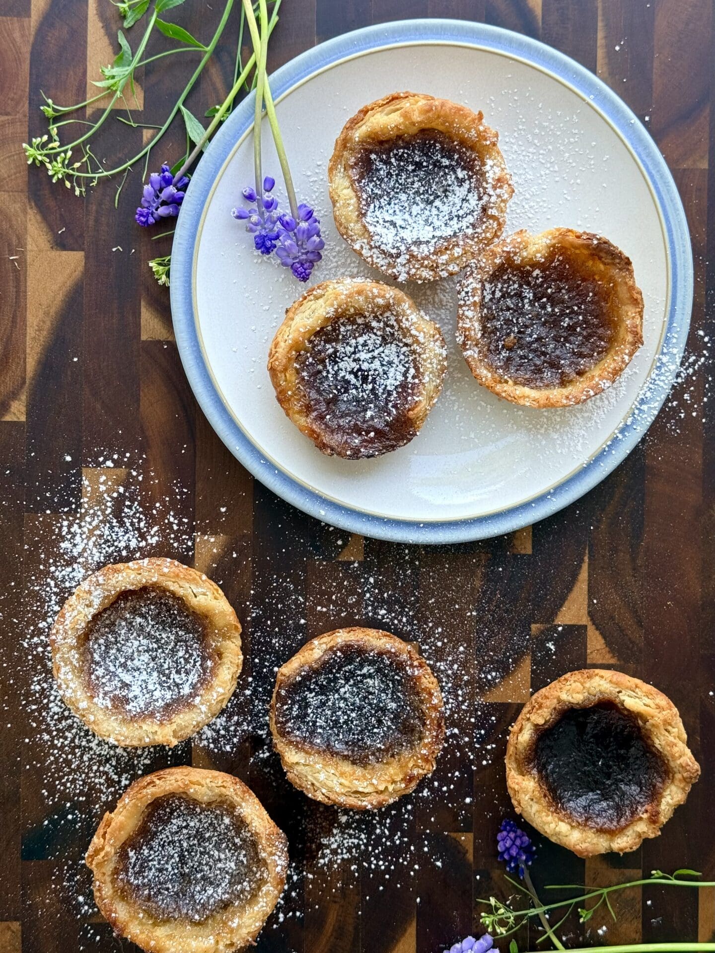 IMG_8440 Freshly baked butter tarts are seen from above. Some sit on a pretty blue and white plate, and some rest directly on a wooden board. The tarts are dusted in confectioners sugar and surrounded by small purple and white flowers.