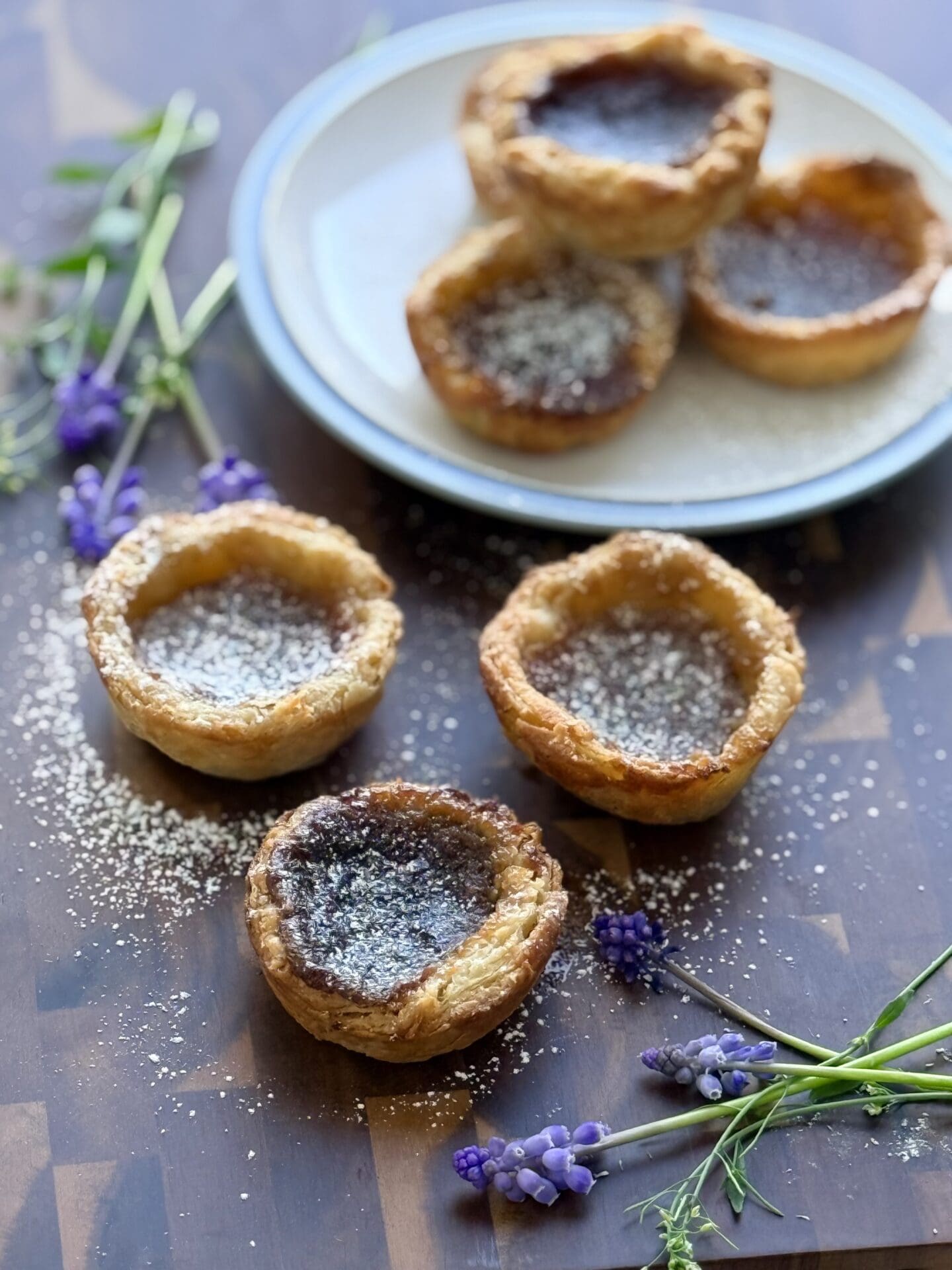 IMG_8458 A trio of butter tarts sits in the foreground of the photo, on a wooden board. In the background, a small blue and white plate holds another 3 tarts. Small, purple flowers complete the scene.