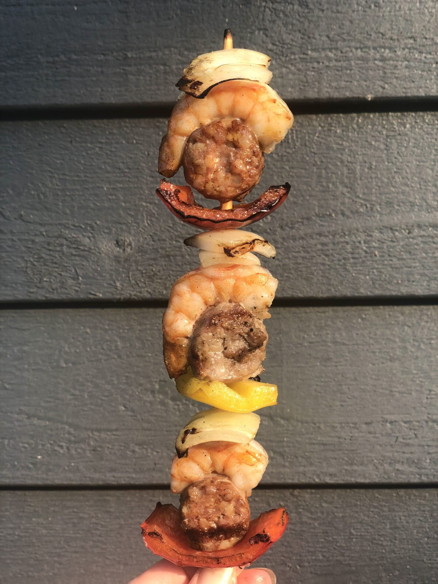 A grilled skewer is held up against the siding of a cottage, showing its repeating pattern of bell peppers, sausage, shrimp and onion.