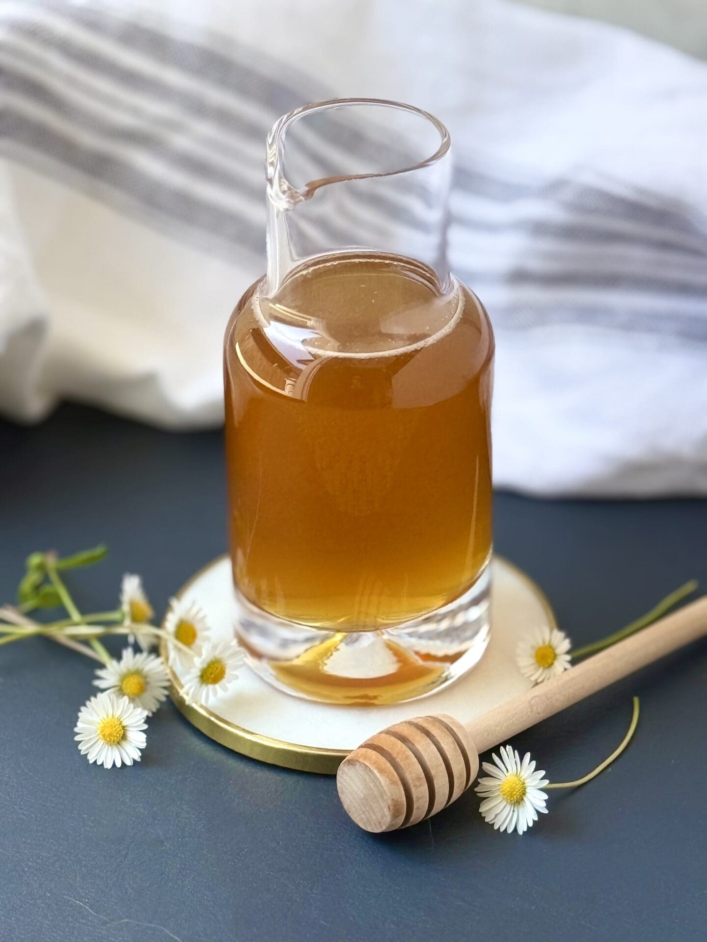 A glass beaker of Honey Simple Syrup sits on a navy blue background, surrounded by tiny daisies and a honey dipper.