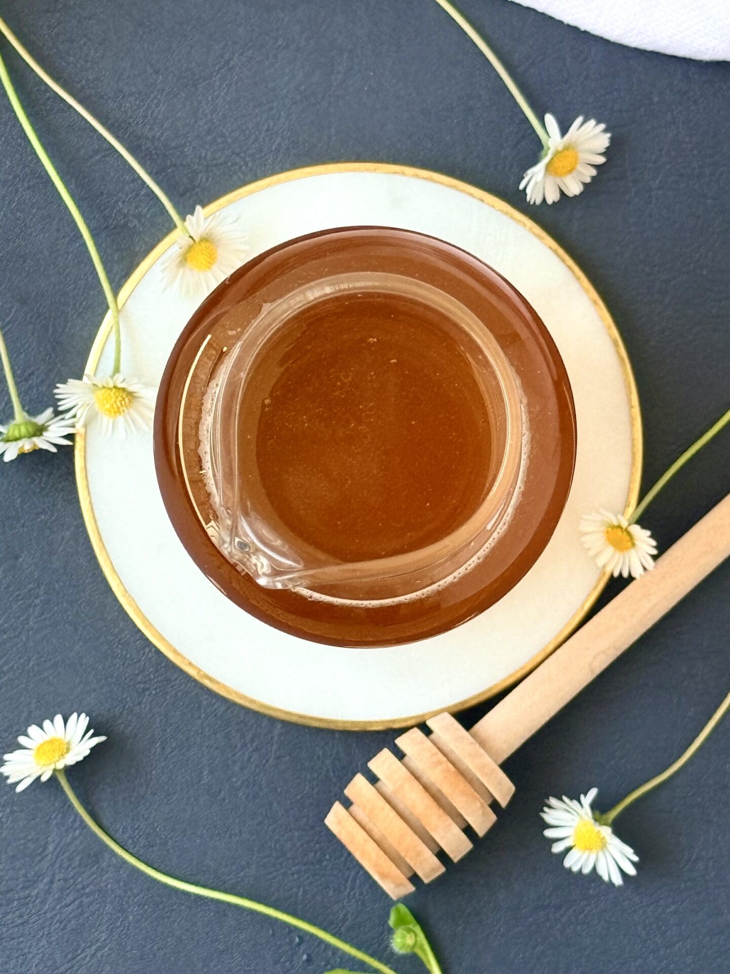 A glass bottle of honey simple syrup is seen from above, resting on a white and gold coaster. Tiny white and yellow daisies and a wooden honey dipper surround the syrup.
