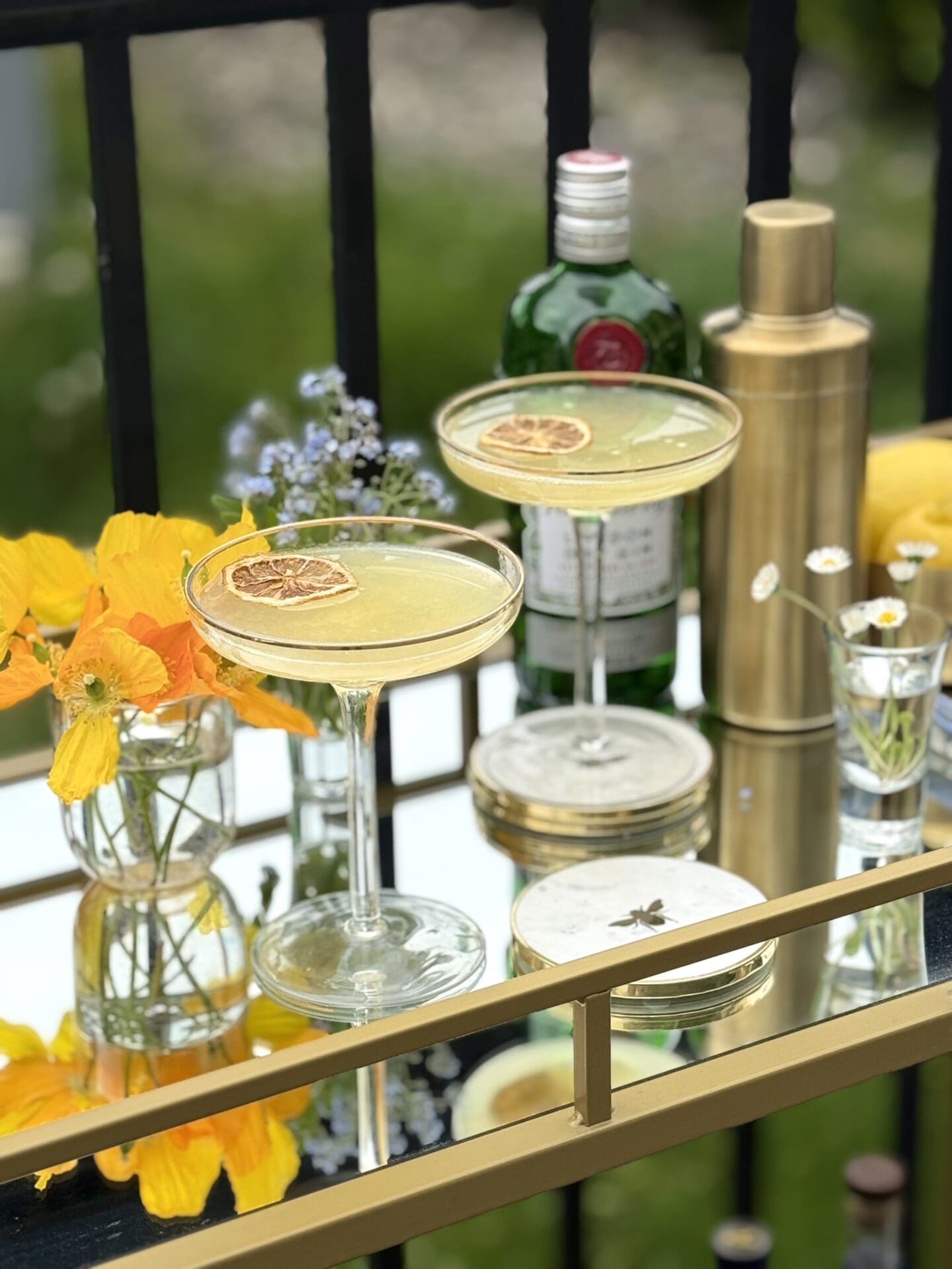 Two coupe glasses filled with Bee’s Knees cocktails sits on a gold and glass bar cart, set outdoors on a warm, sunny day. The cocktails are surrounded by fresh flowers, a gold cocktail shaker and a bottle of gin.