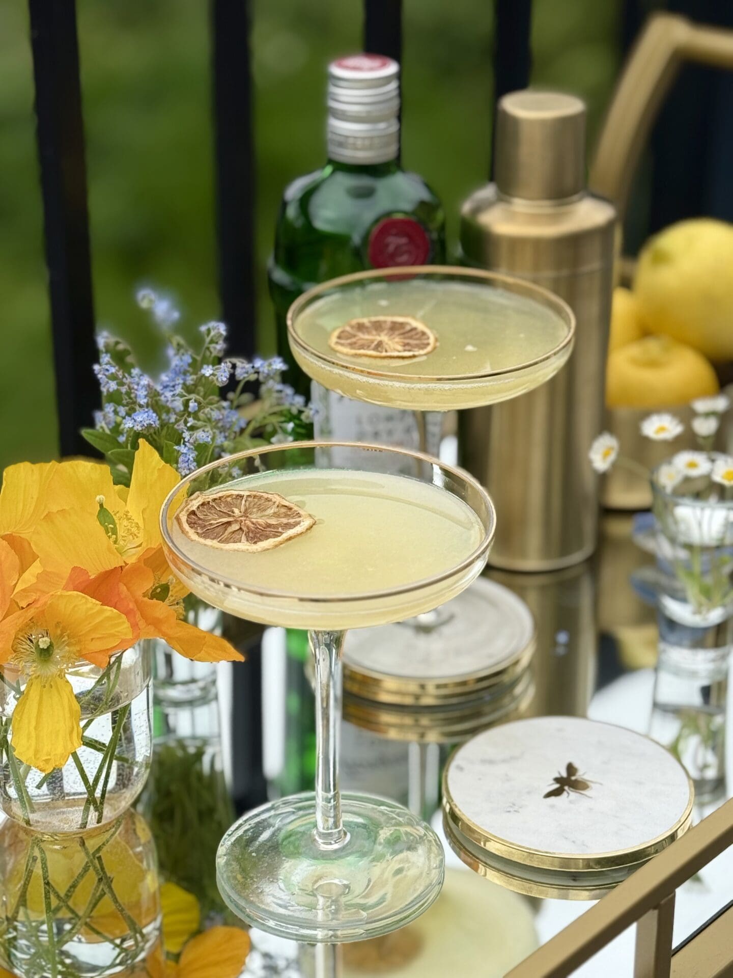 A bar cart filled with the ingredients to make a Bee’s Knees cocktail sits on a sunny patio. Two coupe glasses filled with the drink are garnished with dried lemons slices and ready to be served.