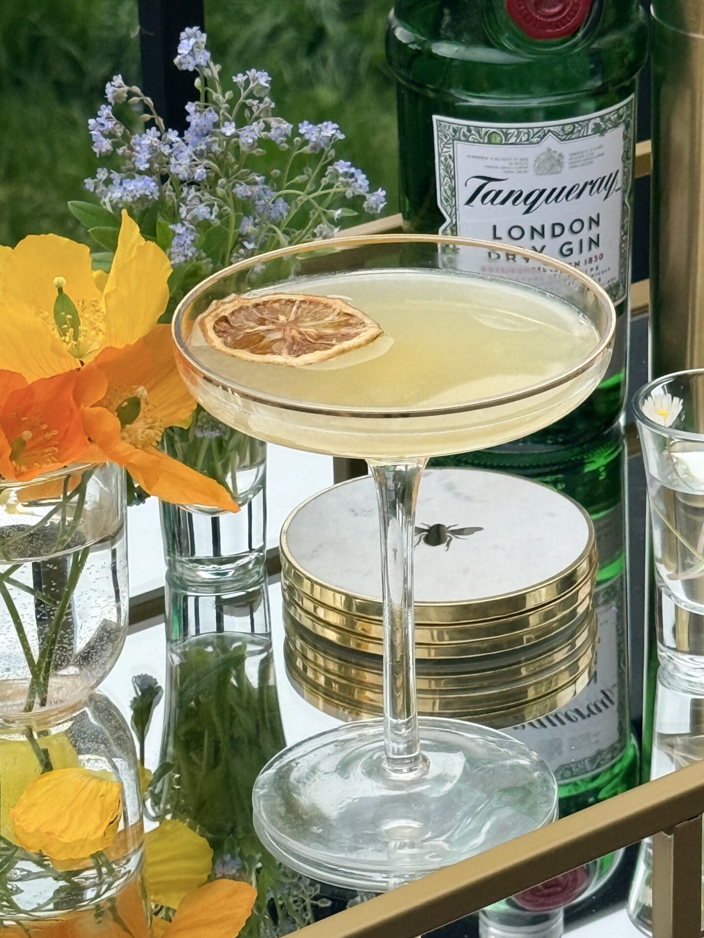 A coupe glass filled with a Bee’s Knees cocktail sits on a gold bar cart, surrounded by fresh flowers. A bottle of gin can be seen in the background.