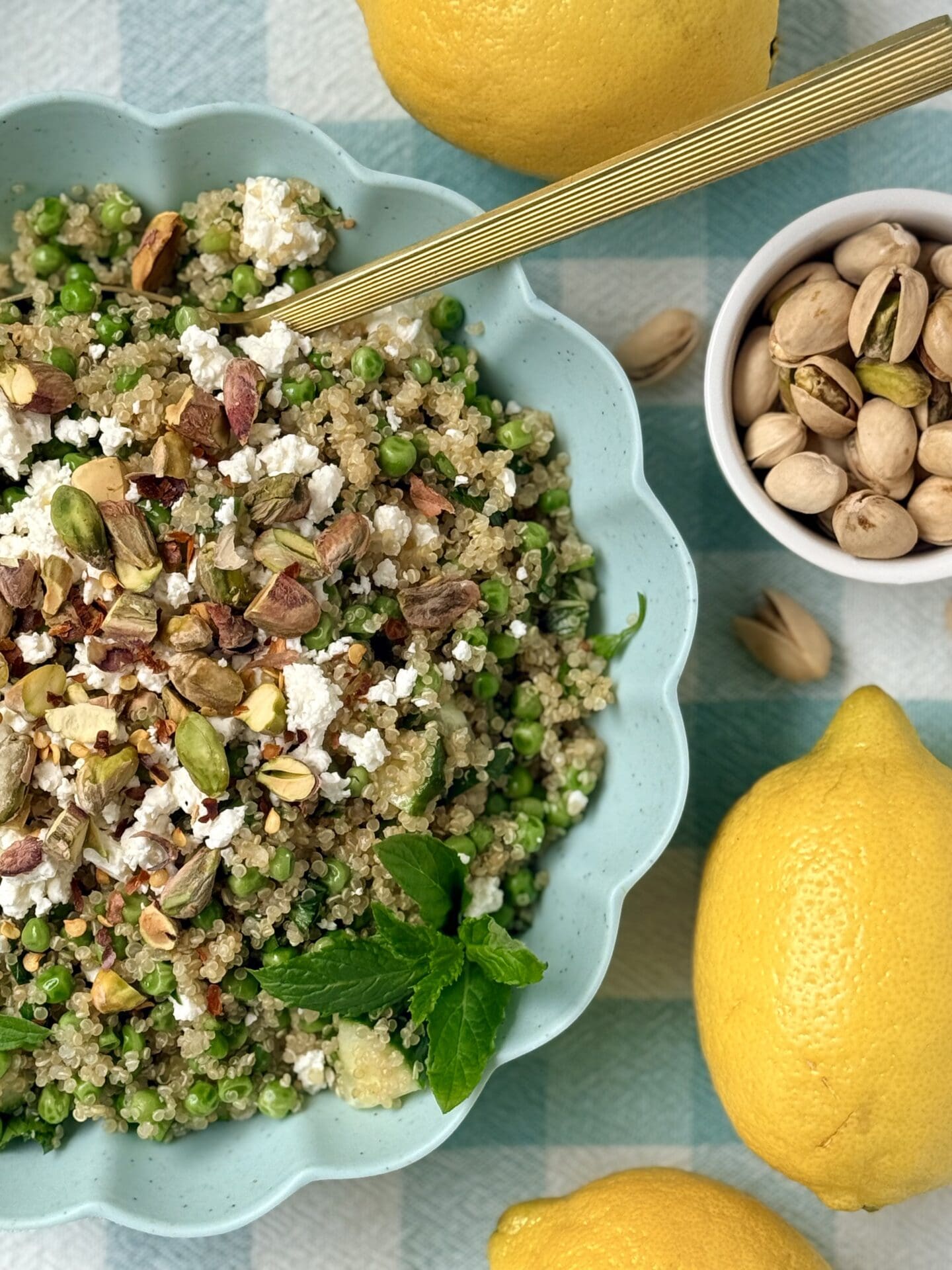A turquoise bowl with a scalloped edge is filled with Lemony Quinoa Salad with Spring Peas and Pistachios. The bowl rests on a checkered tablecloth and is surrounded by pistachios and lemons.