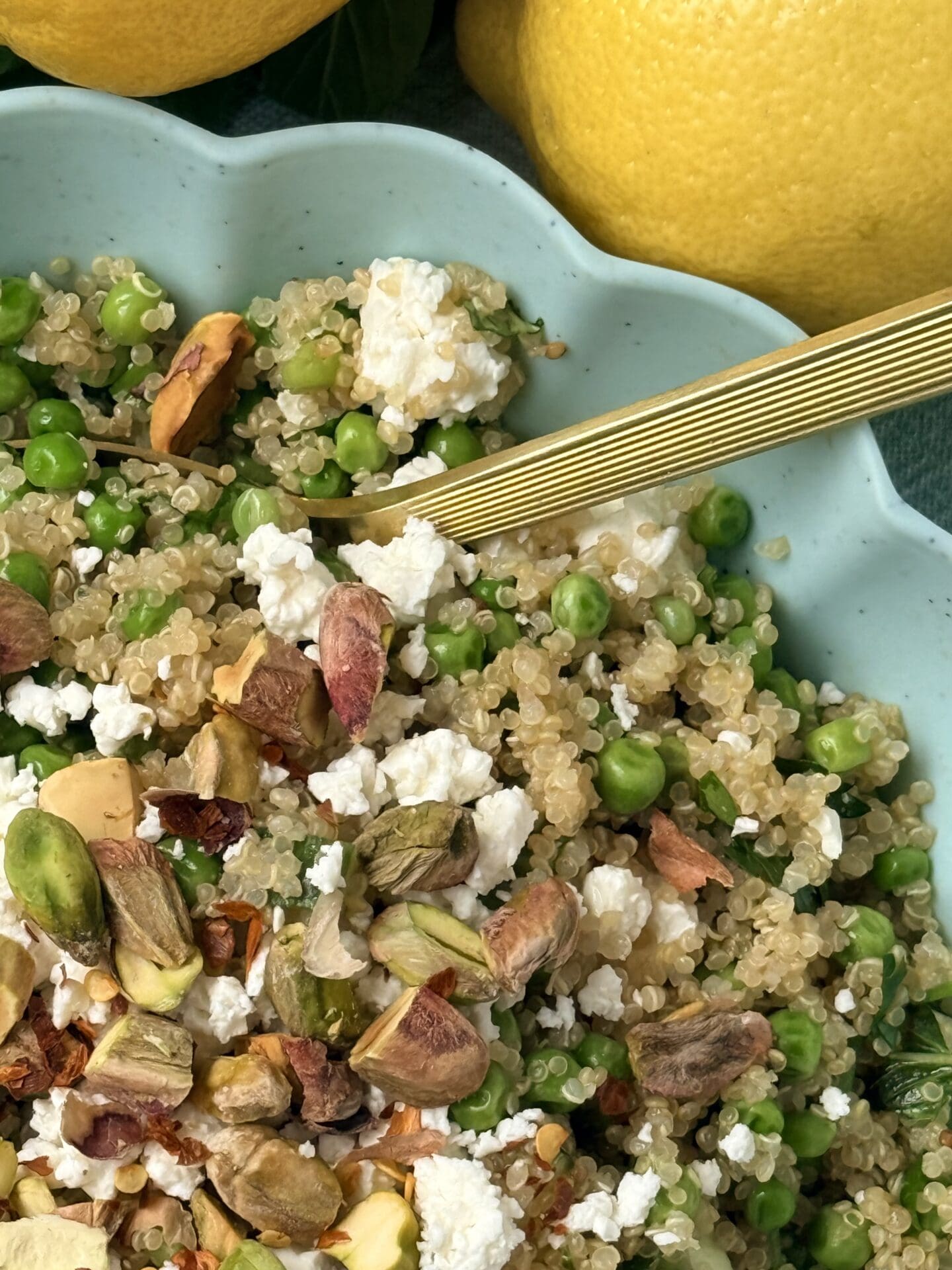 A quarter of a bowl of Lemony Quinoa Salad with Spring Peas and Pistachios is seen from above with a gold serving spoon and fresh lemons.
