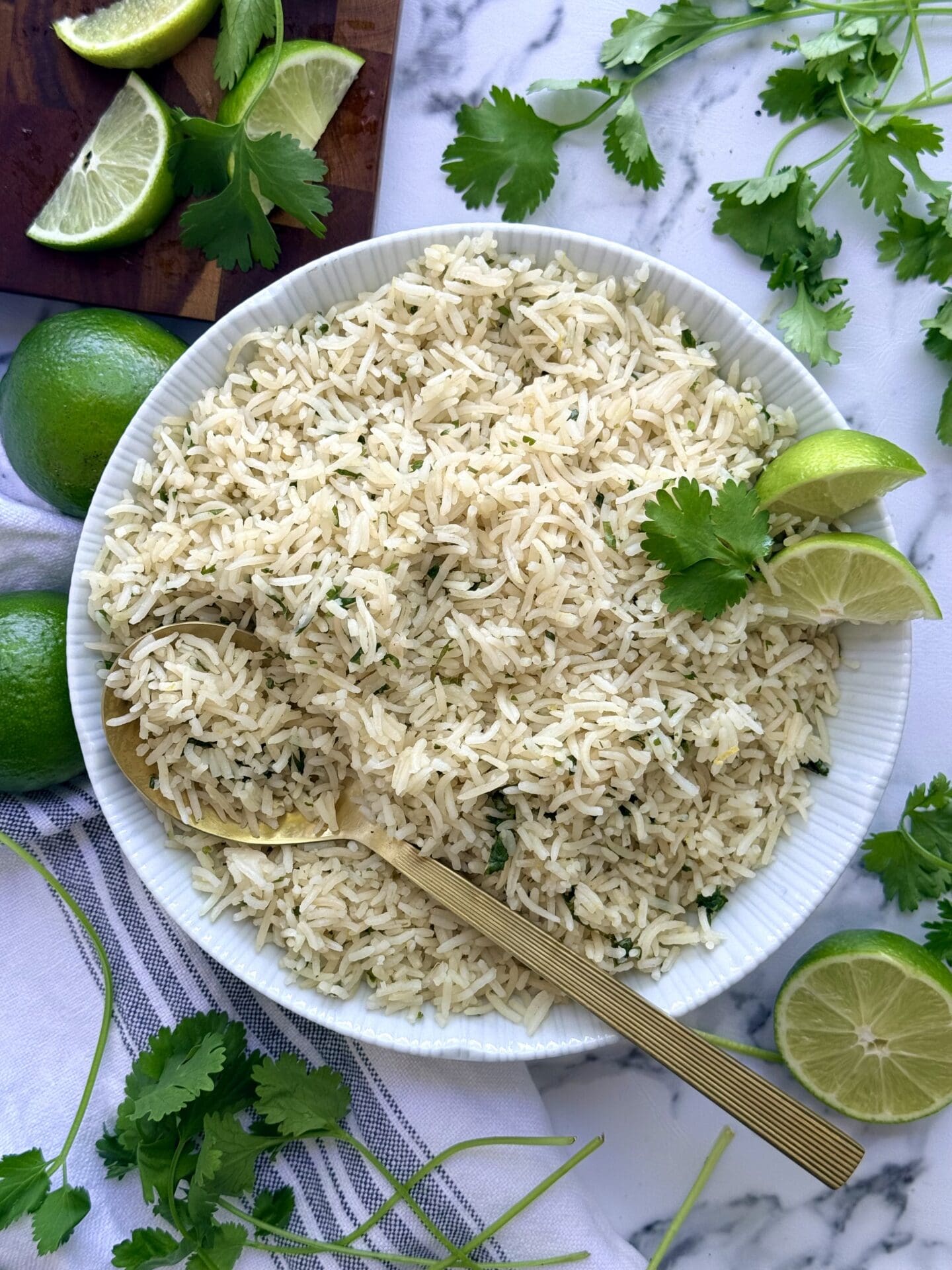 IMG_8962 A large bowl of cilantro lime rice sits in the centre of the frame. The bowl rests on a white marble counter top. A gold coloured serving spoon is seen scooping a large serving. The rice is surrounded by fresh lime slices and cilantro leaves.